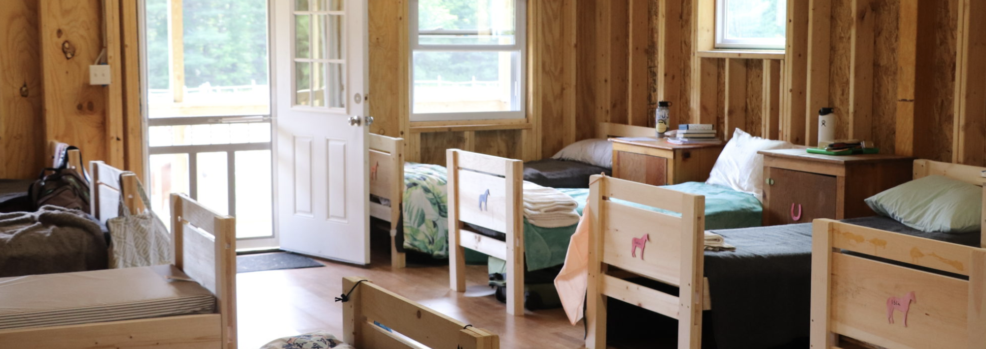 Interior of a camp cabin with multiple wooden bunk beds, open door, and natural wood paneling.