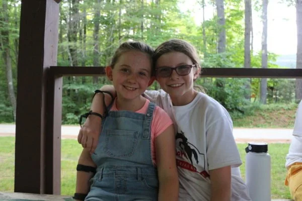 Two smiling girls sitting together on a wooden bench in an outdoor setting. One girl is wearing overalls and the other is in a t-shirt, with trees visible in the background.