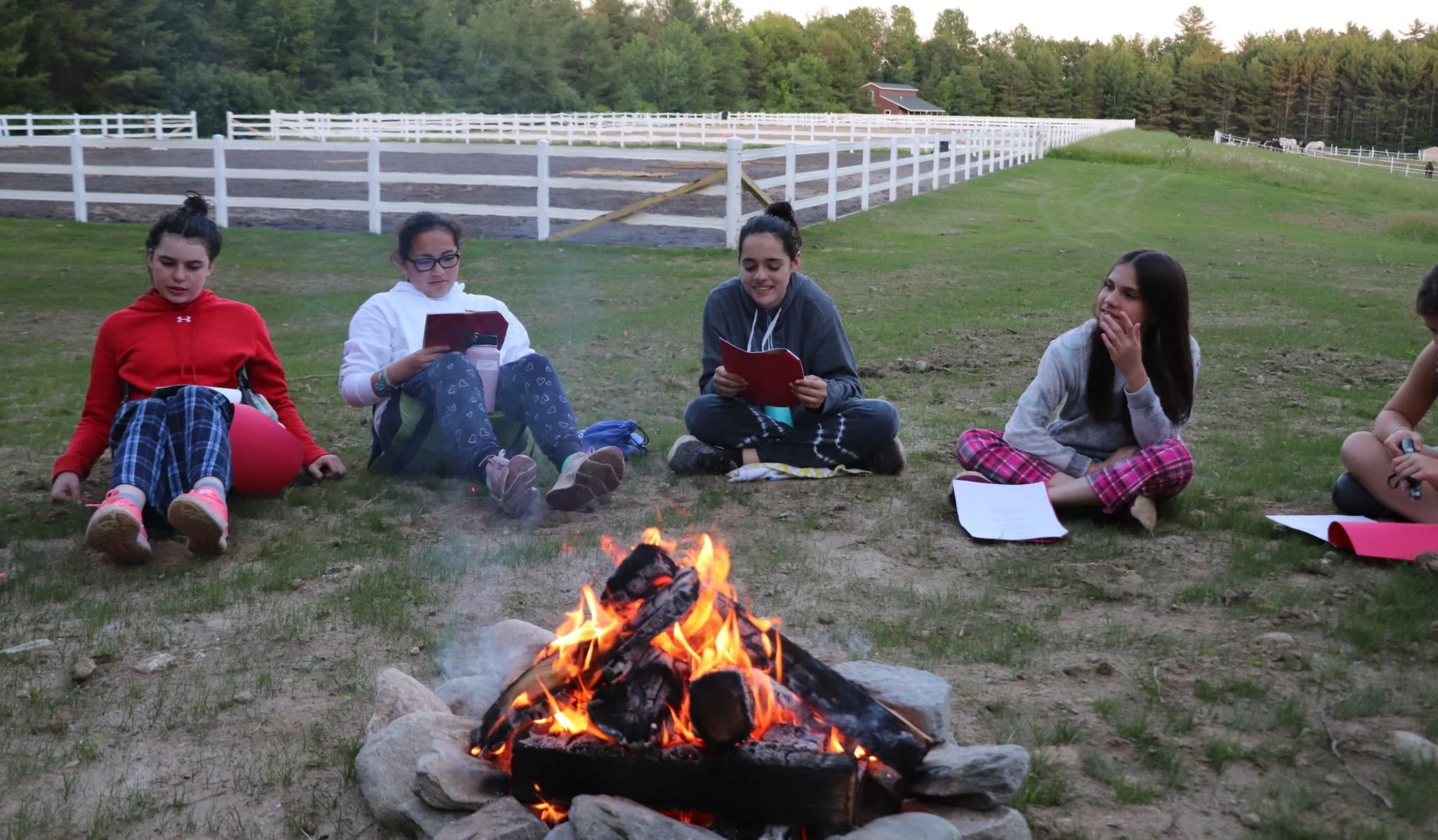 Group of people sitting around a campfire in a field, holding notebooks, with white fencing and trees in the background.