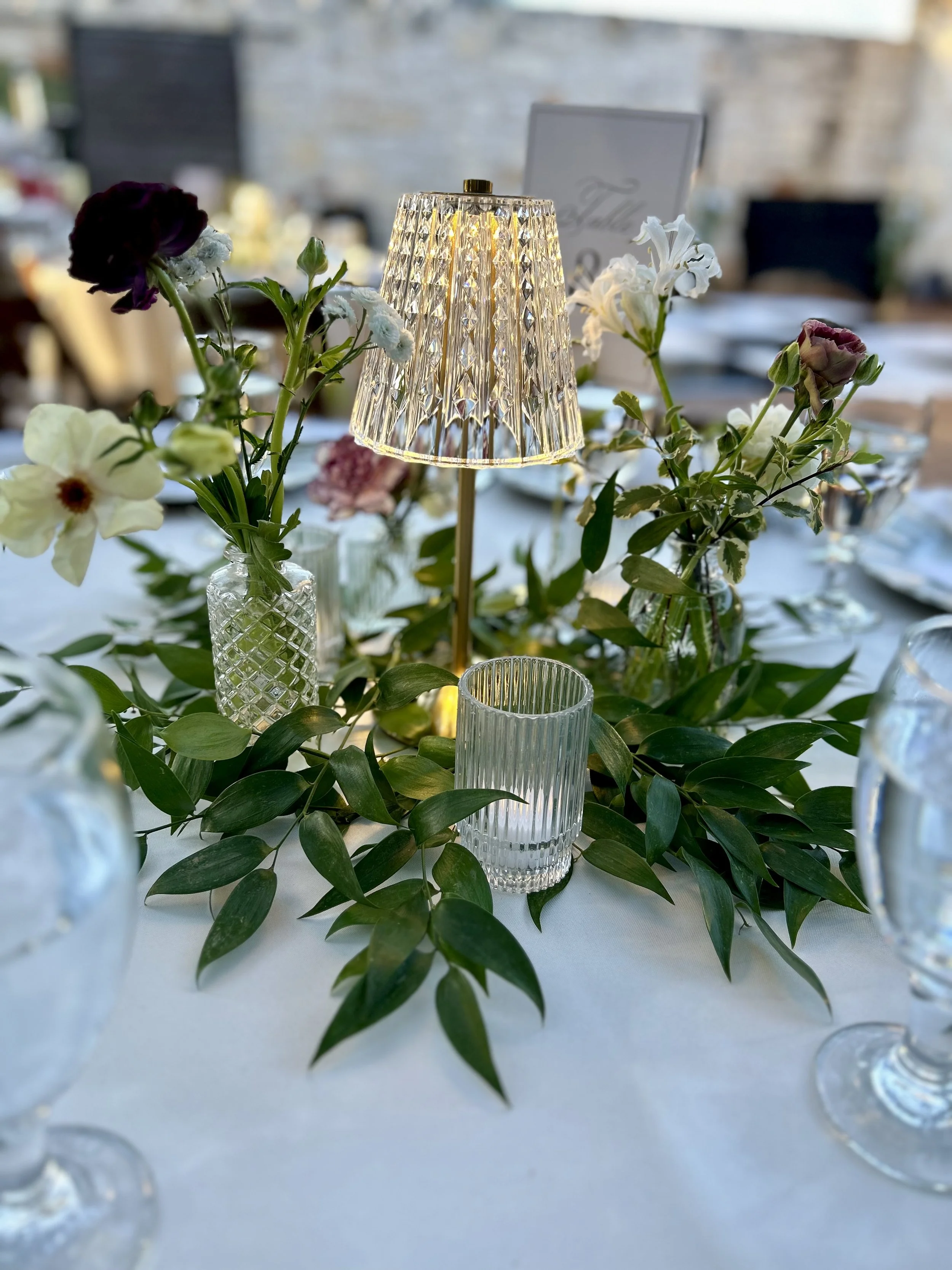 Reception table decor with greenery, votive candles, and small glass lamp