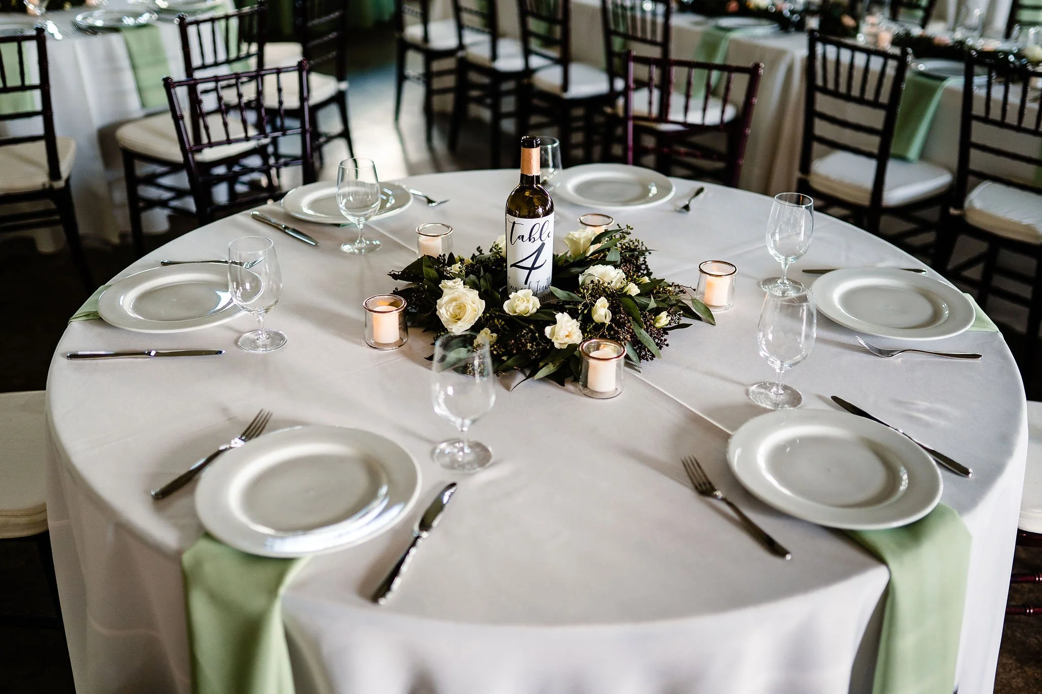 Round Guest table with white plate, sage cloth napkins draping down and a wine bottle table number with greenery at the Vineyard at Florence Texas