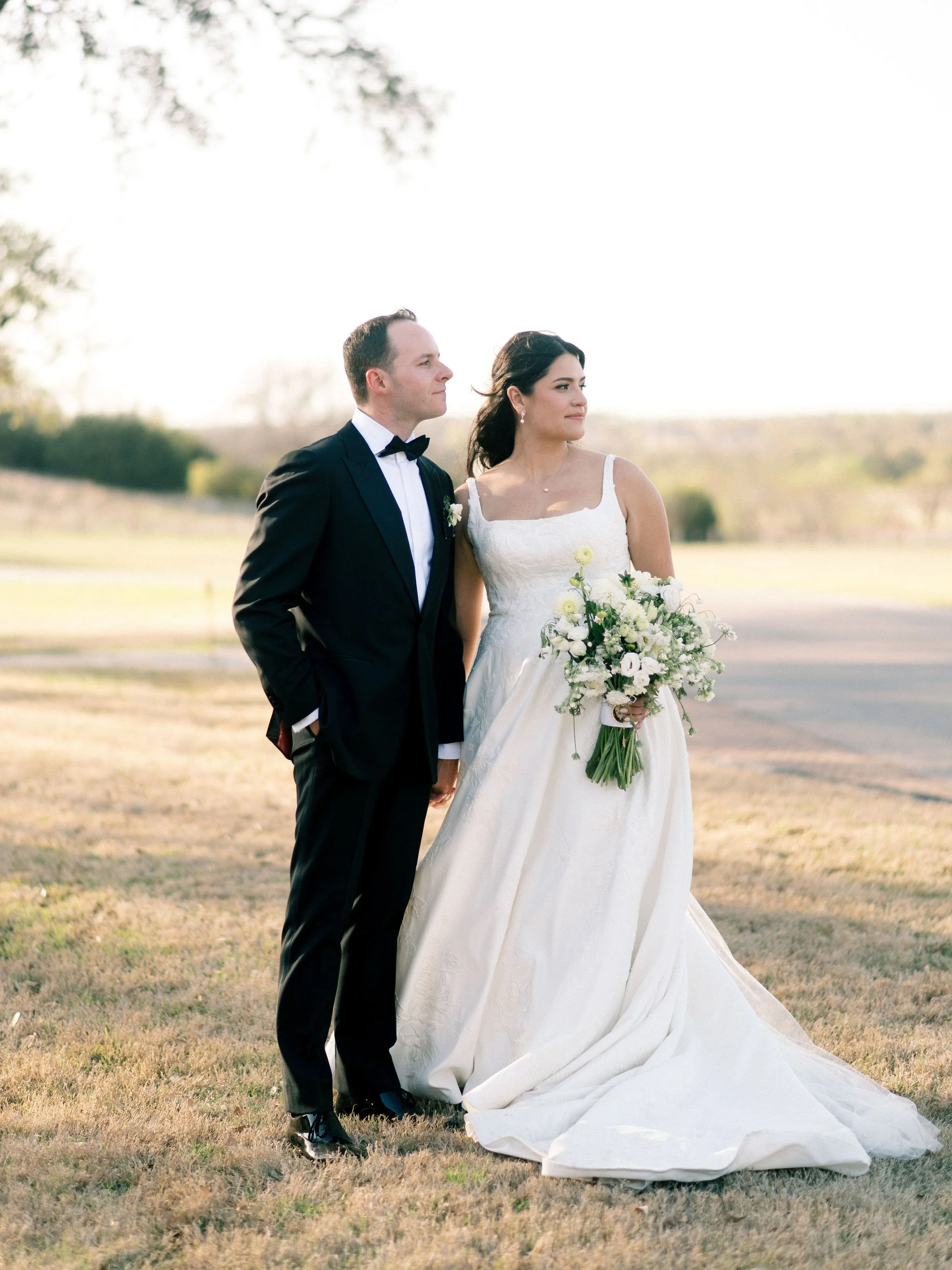 A bride and groom standing outdoors on their wedding day, with the bride holding a bouquet of white flowers and wearing a white wedding gown, and the groom in a black tuxedo with a bow tie, both looking to the right.