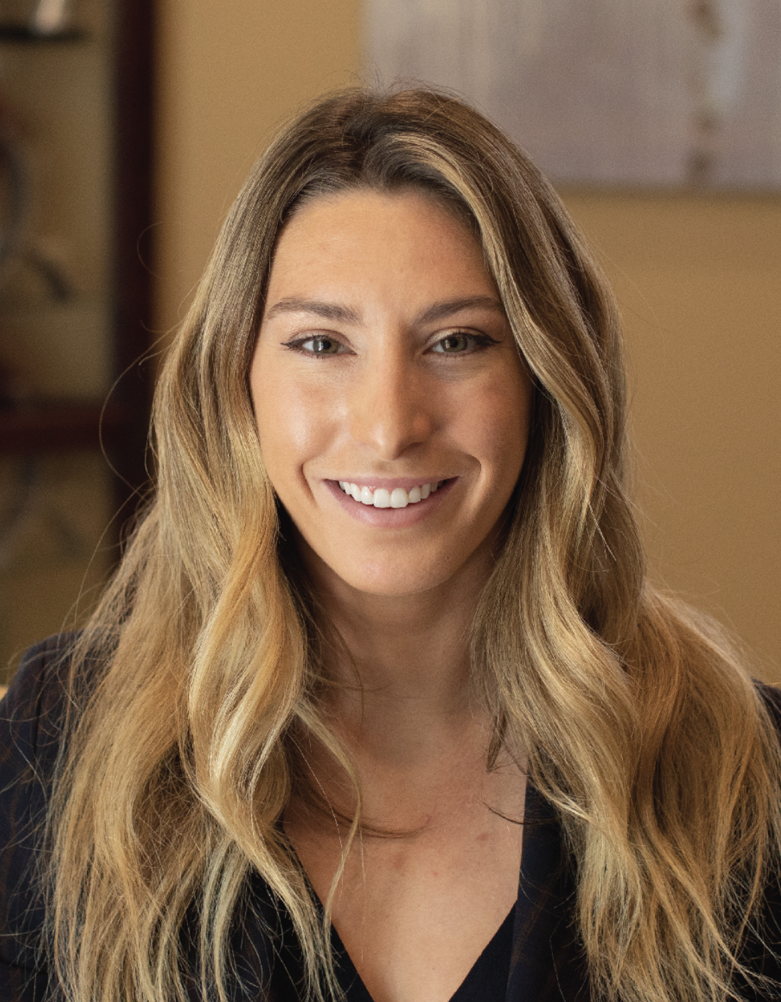 Smiling woman with long wavy hair, wearing a dark top, indoors.