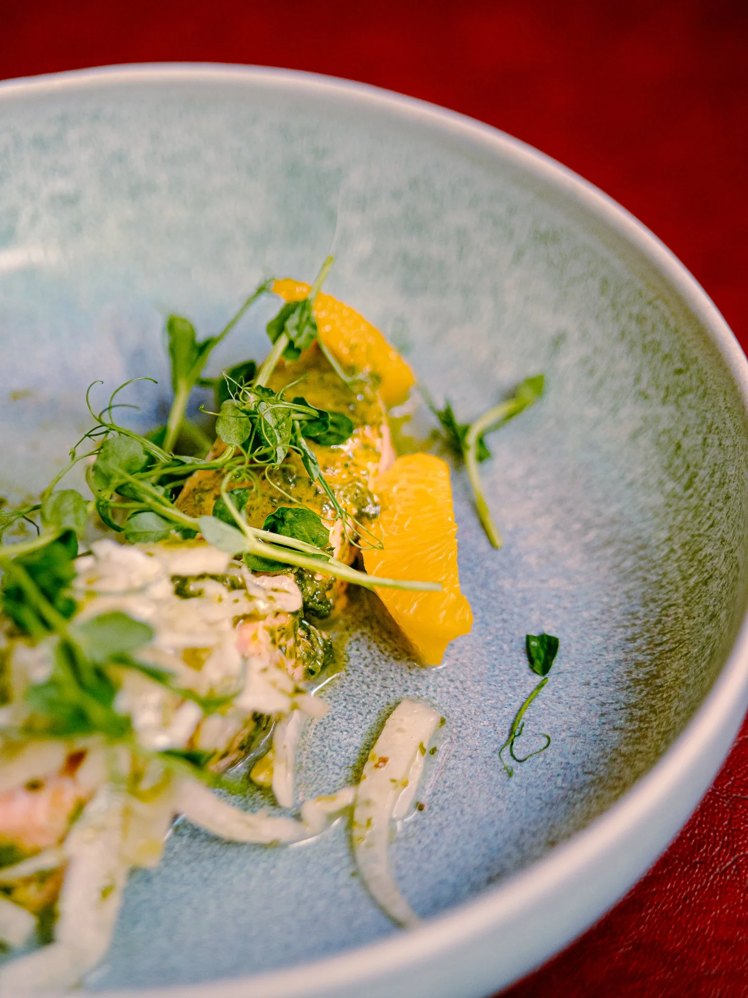 Une assiette de carpaccio de poisson garnie de micro-pousses, rondelles de citron, et crème, sur un plat bleu foncé.