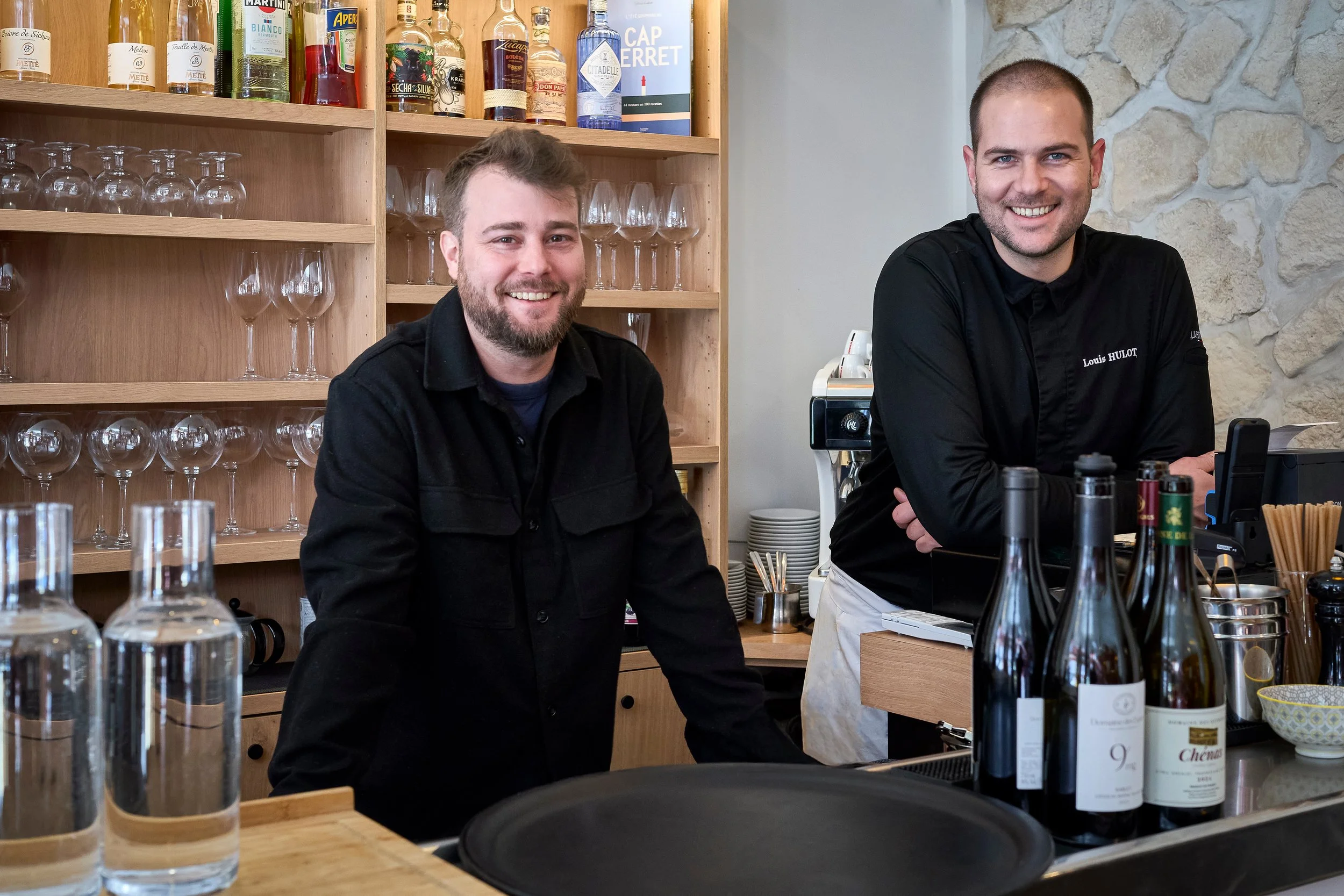 Deux hommes souriants derrière un bar avec des bouteilles d'alcool, des verres et des accessoires de bar, dans un décor en pierre.