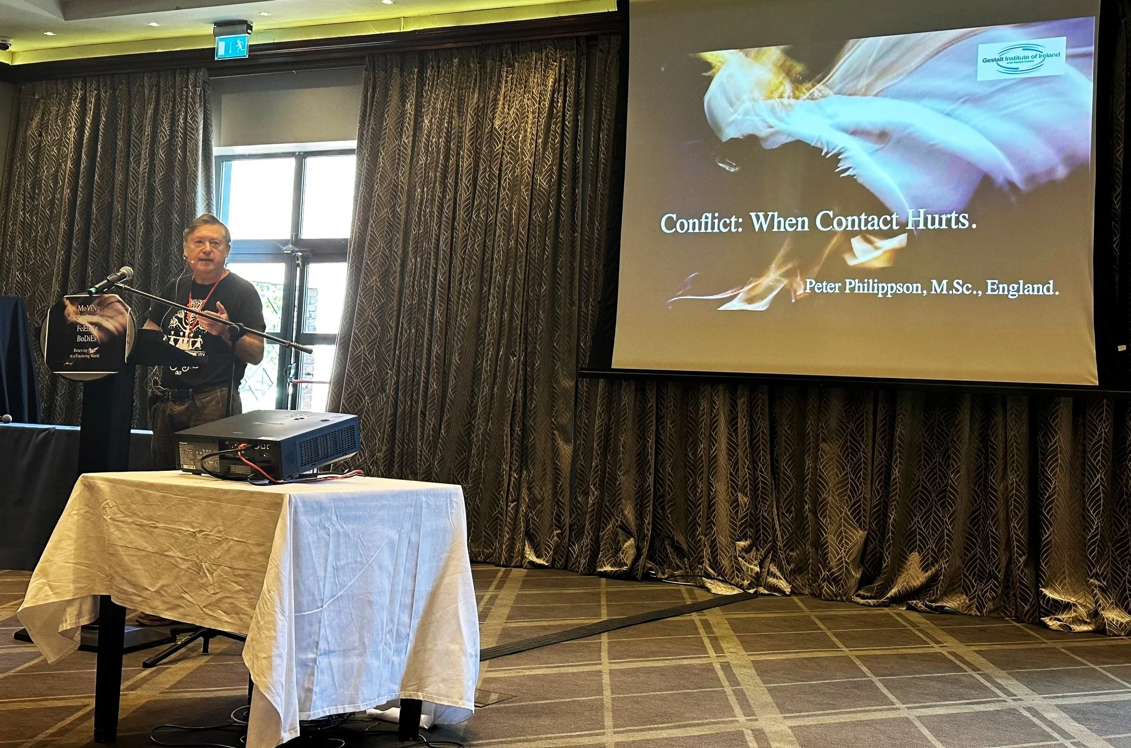 A person speaking at a conference or seminar, standing at a podium with a microphone. A large screen displays a presentation slide titled 'Conflict: When Contact Hurts' by Peter Philippson, M.Sc., England. The room has patterned curtains and a table
