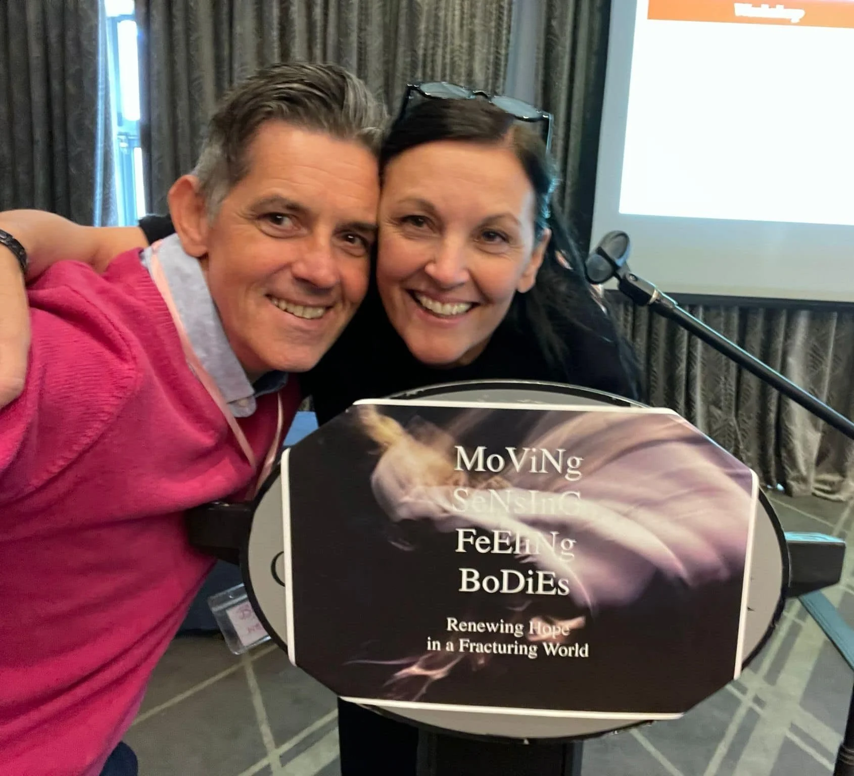 Two smiling people leaning into each other at a presentation or conference, holding a sign with the text 'Moving Sensing Feeling Bodies: Renewing Hope in a Fracturing World'.