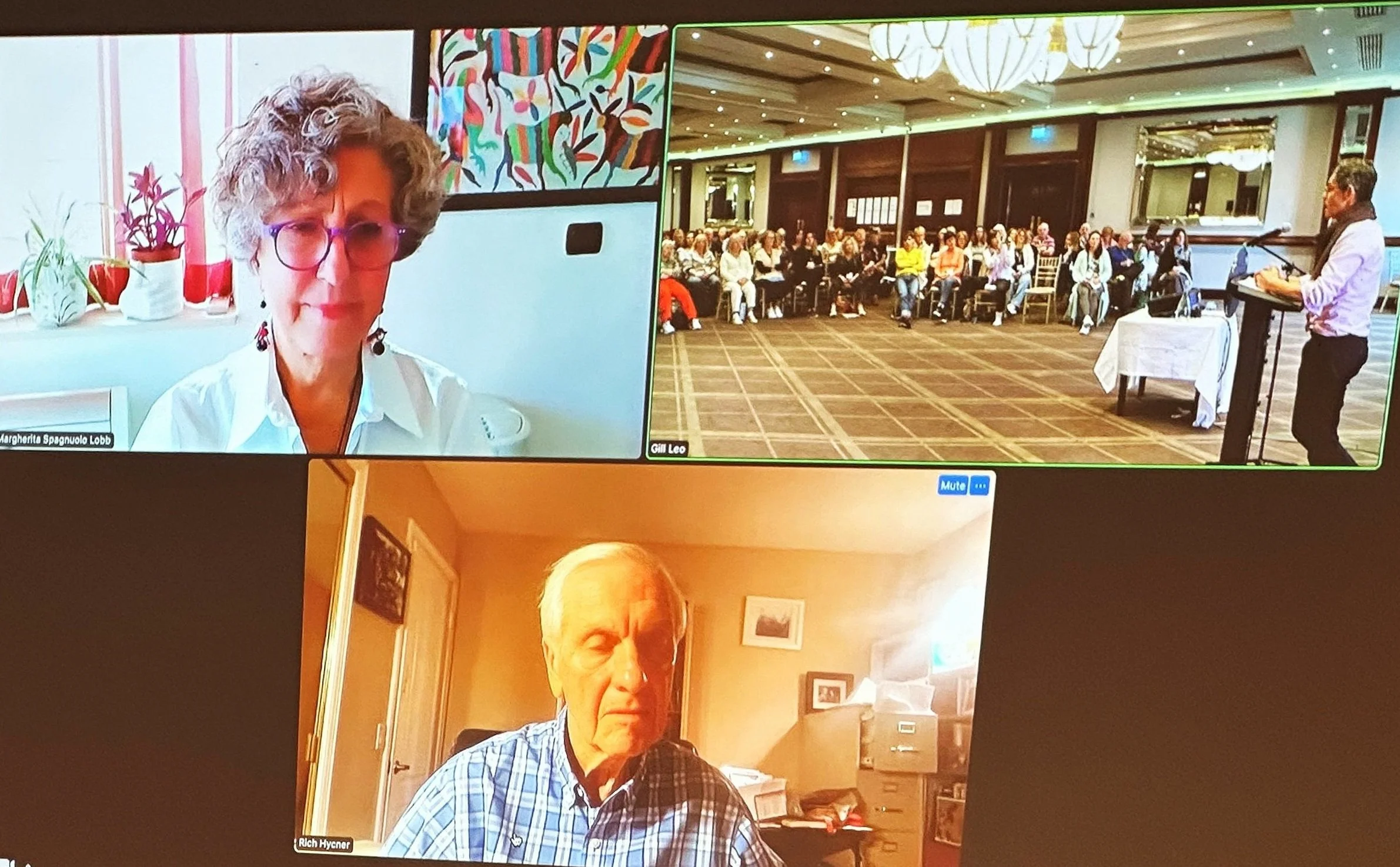 Screenshot of a virtual meeting with three participants. The top left shows a woman with curly hair and glasses in a white shirt, the top right shows a large audience in a conference room with a speaker at a podium, and the bottom shows an older man