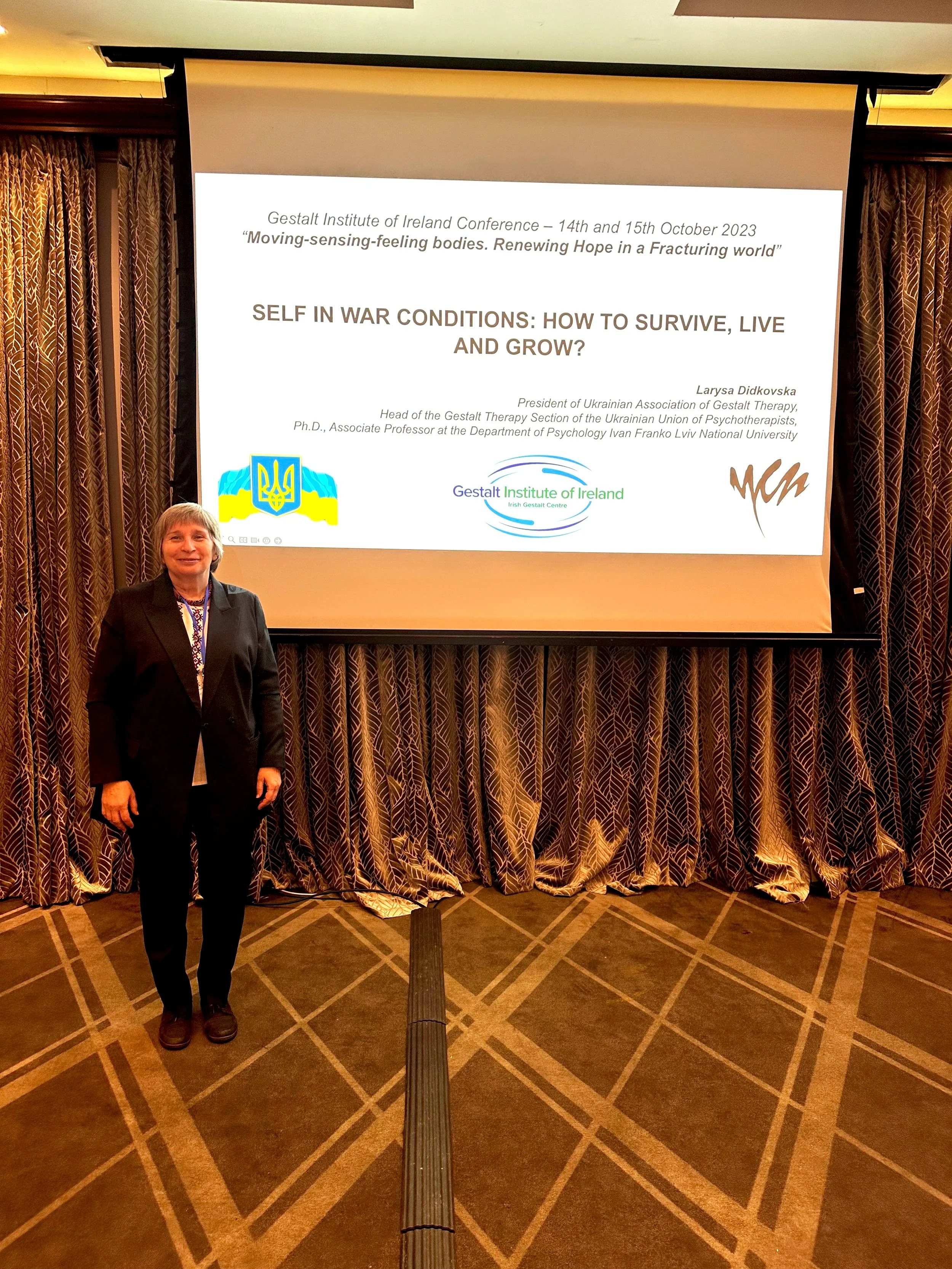 A woman standing in front of a projection screen at a conference. The screen displays a presentation slide for the Gestalt Institute of Ireland Conference, titled 'Self in War Conditions: How to Survive, Live and Grow,' presented by Larysa Didkovska.