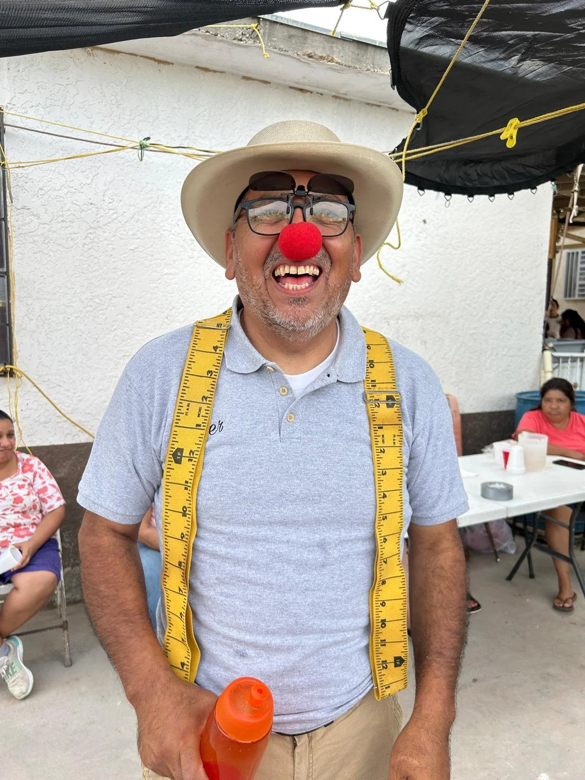 A man dressed as a clown with a red nose, glasses, and a hat, smiling and holding an orange water bottle. He is wearing a yellow measuring tape around his neck and appears to be at a festive outdoor gathering with other people in the background.