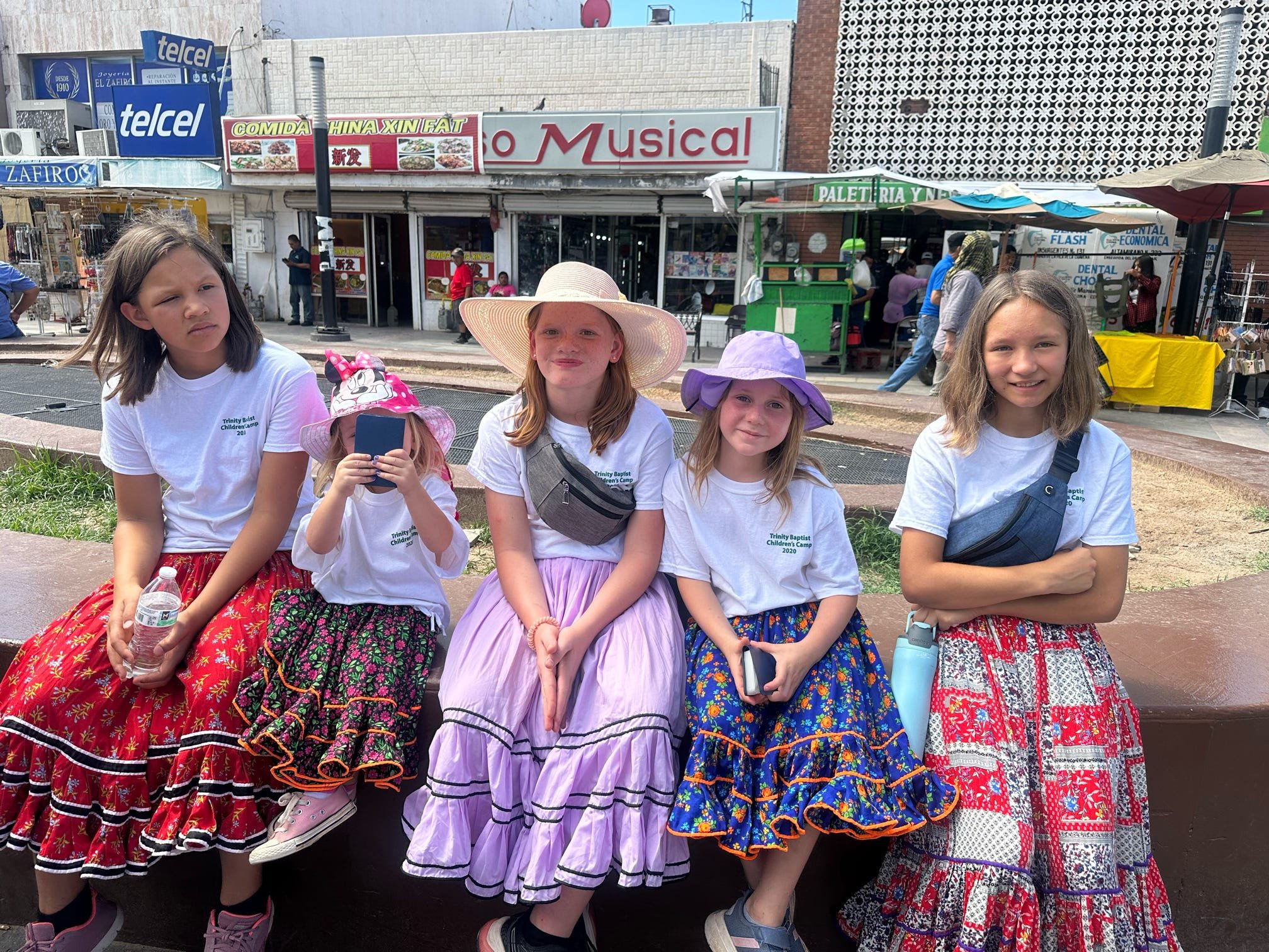 Five young girls sitting on a ledge in traditional Mexican dresses, some wearing hats, outdoors in a busy street area with shops and food stalls visible in the background.