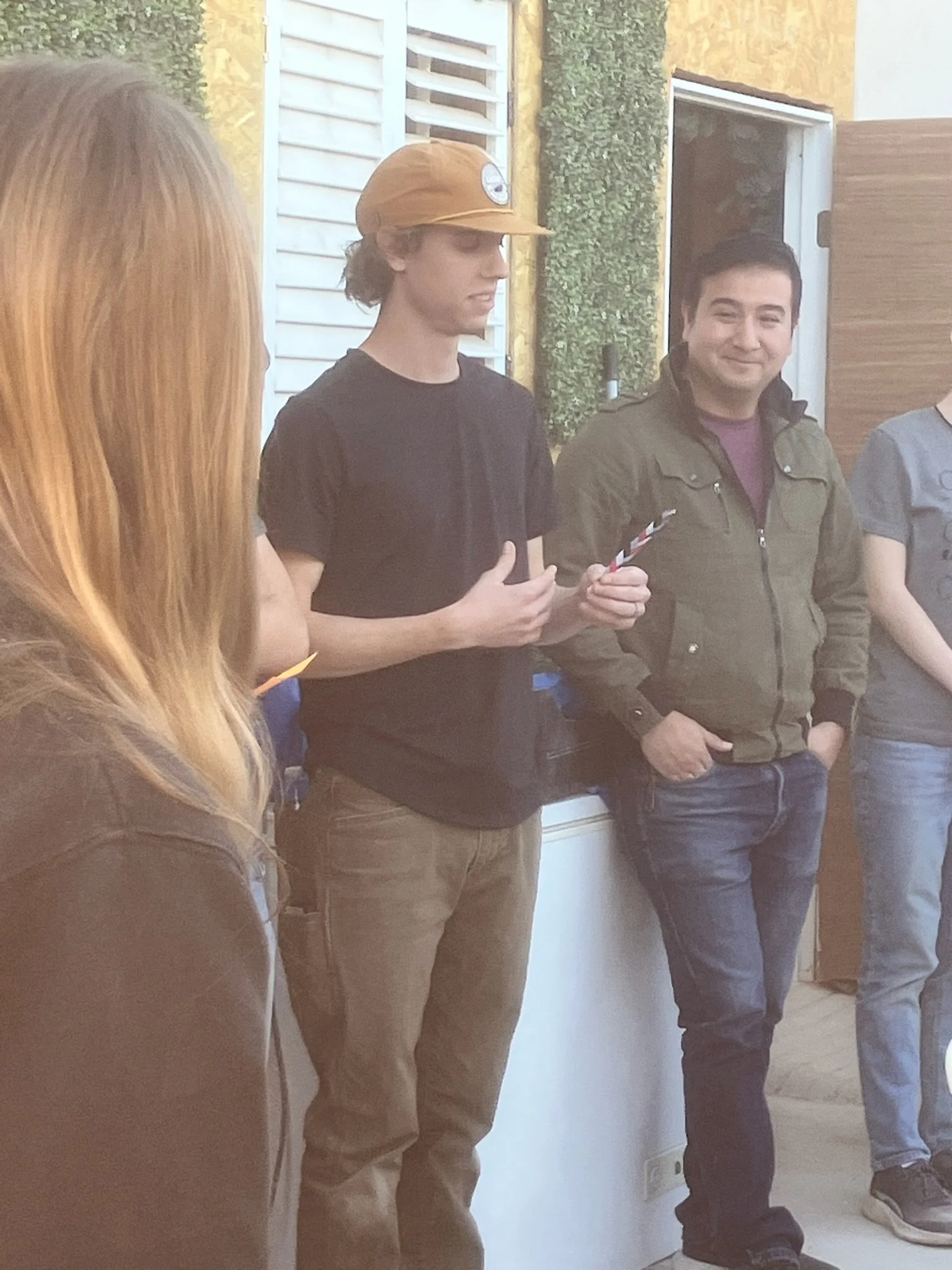 Group of people standing indoors, with one person holding a small striped stick, in a room with white and yellow walls and an open door.