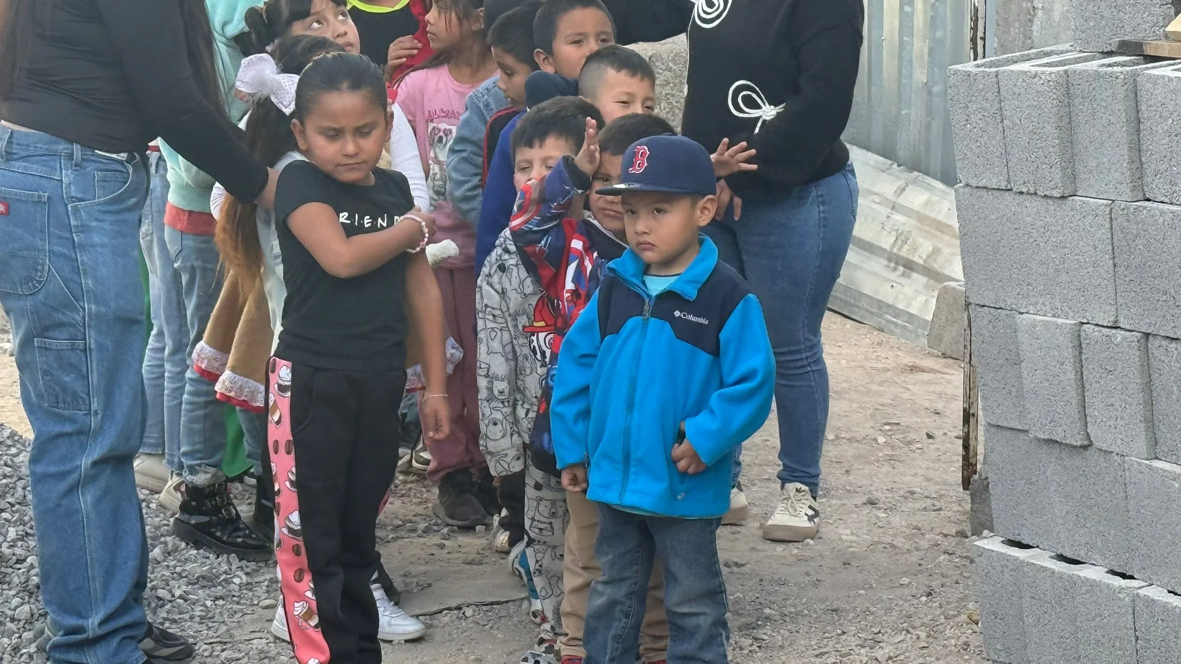 A line of children at a construction site, with some adults nearby, standing on dirt and gravel, facing a stack of cinder blocks.