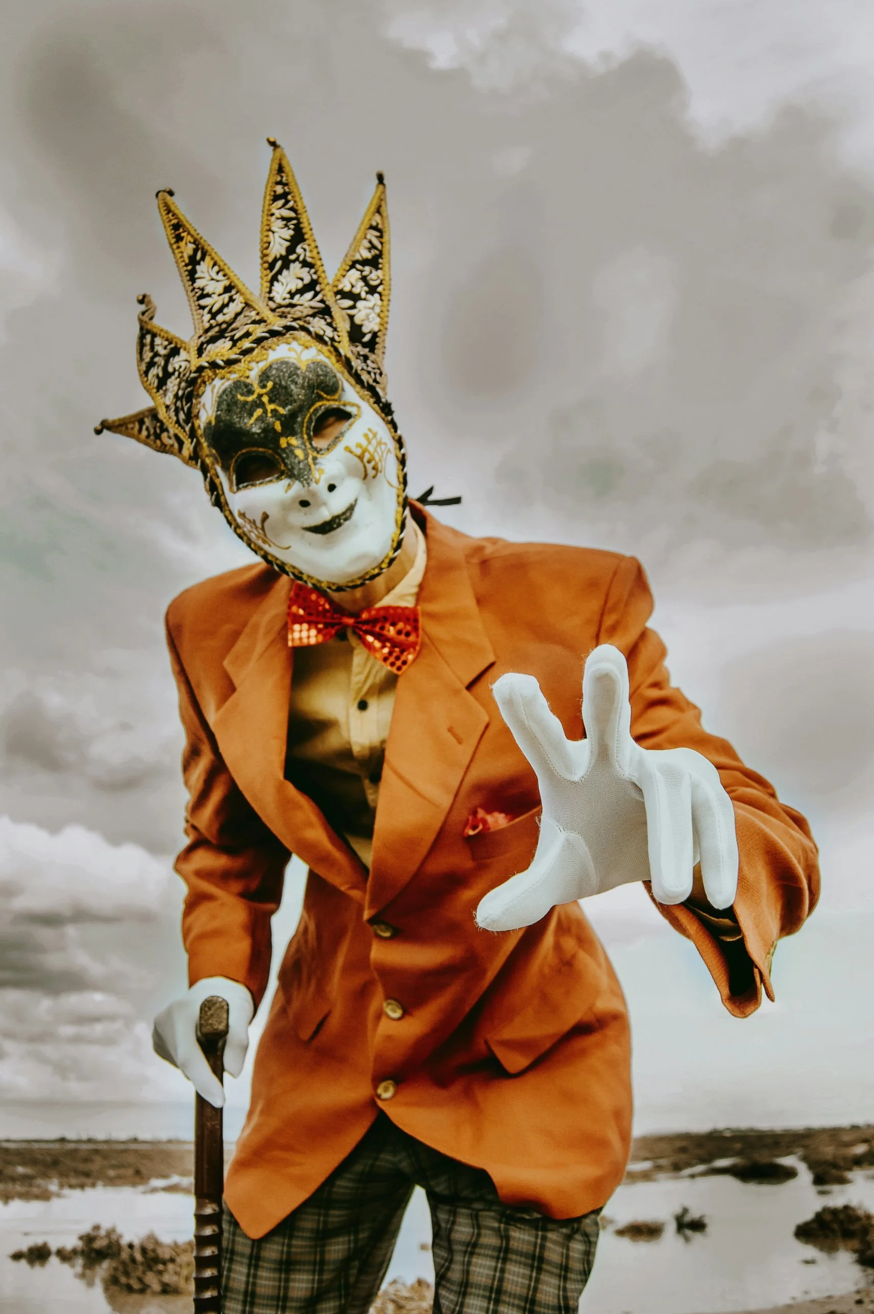 Personnage en veste orange, nœud papillon rouge et gants blancs, portant un masque vénitien de bouffon, appuyé sur une canne et tendant la main vers l'objectif sous un ciel nuageux