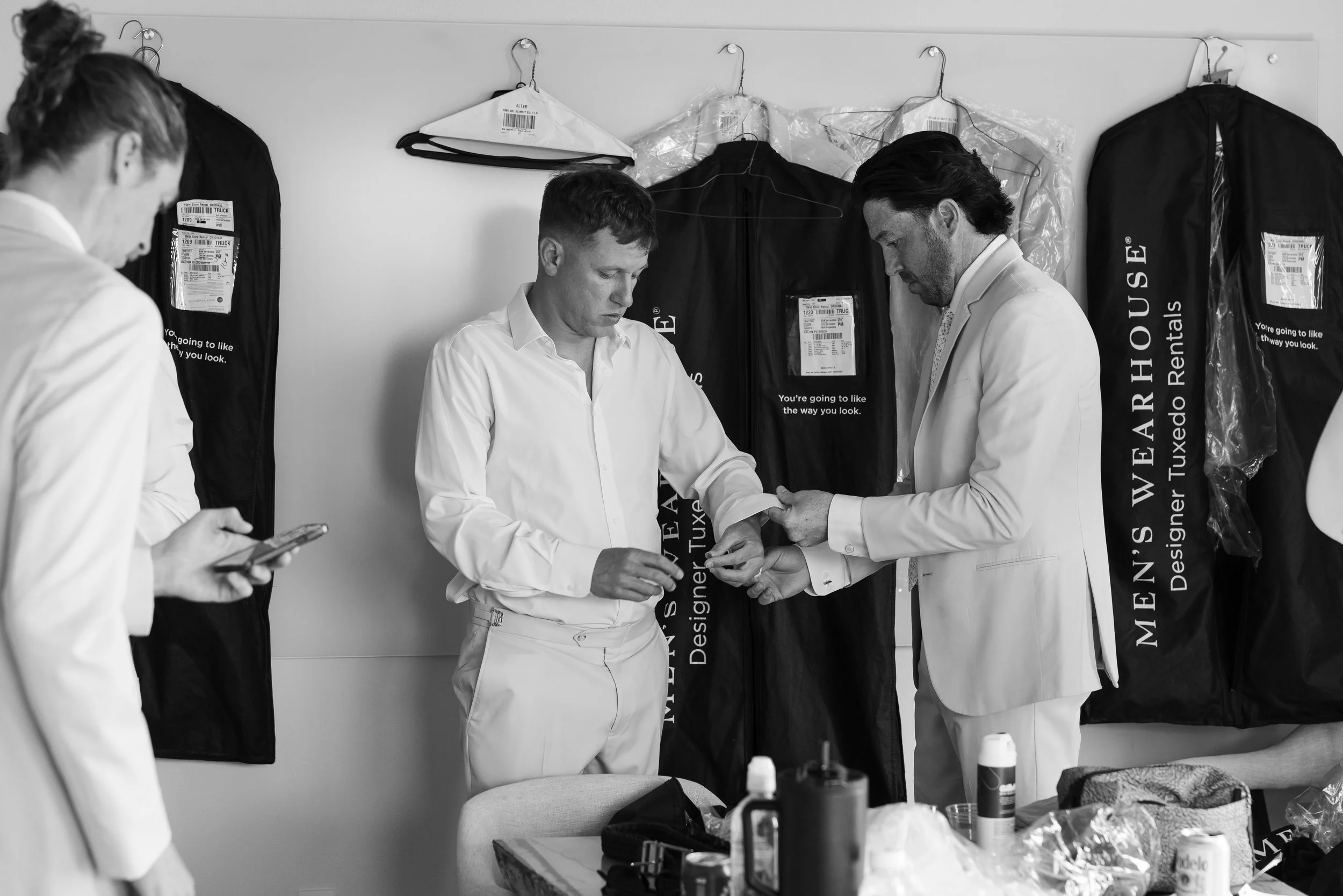 Three men in white suits with black hair, standing in a room with black garment bags hanging on the wall, some in plastic protective covers. The men are preparing or inspecting something, with one man on the left using a phone, and the other two look
