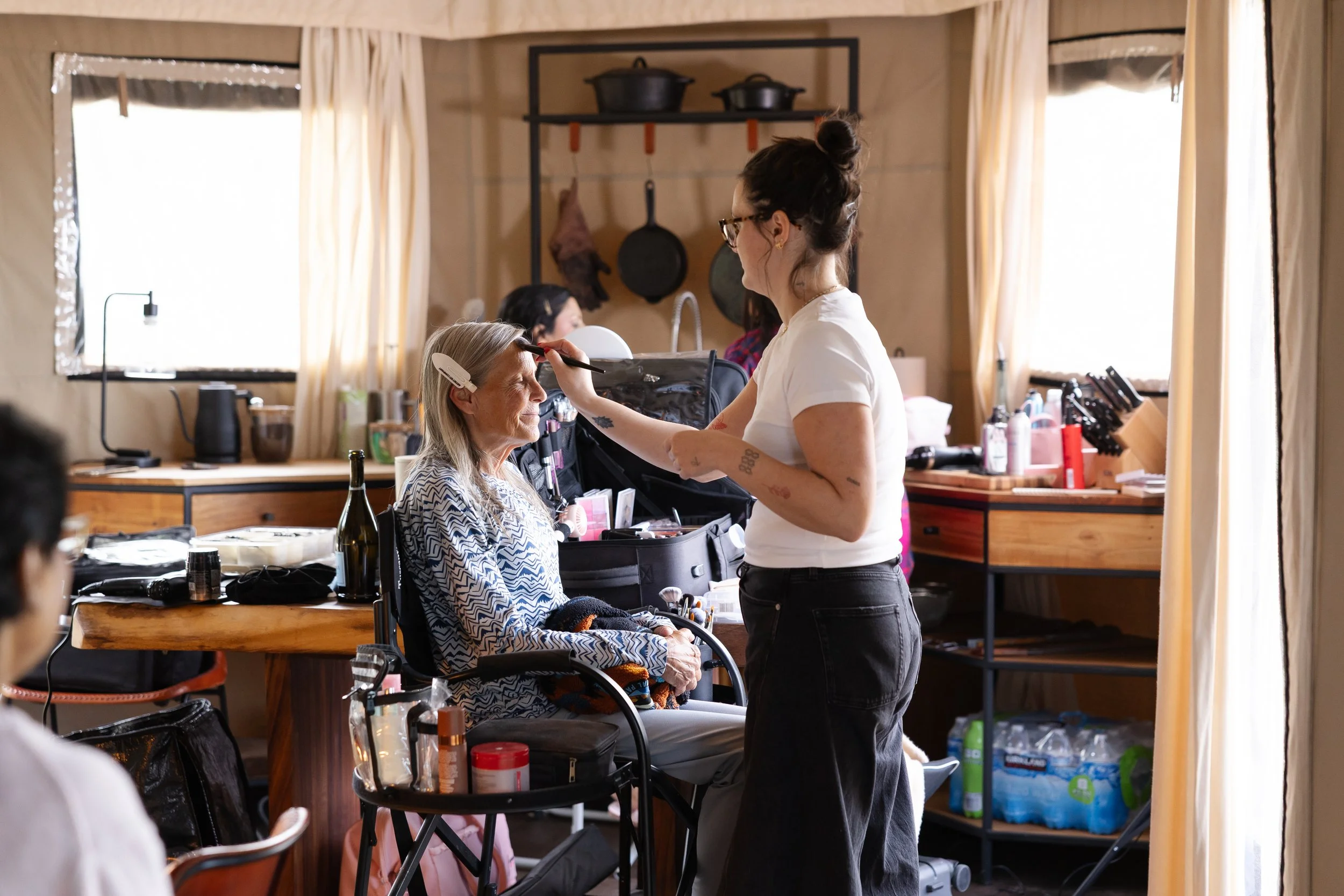 A makeup artist applying makeup to an elderly woman sitting in a chair at a table with makeup products, in a cozy room with large windows and curtains at Walden Retreat.