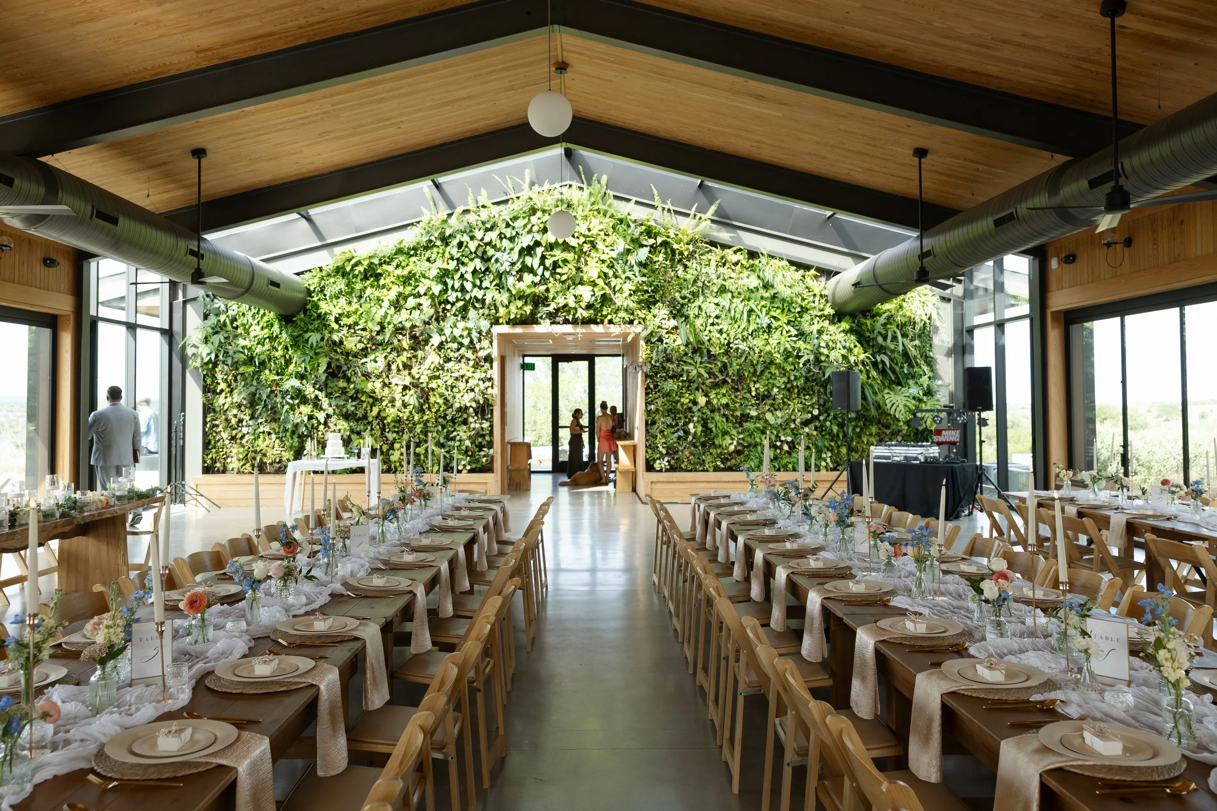 Interior of a wedding venue with long wooden tables decorated with flower arrangements, set for a celebration, and a green wall archway at the entrance in the reception space at Walden Retreats.