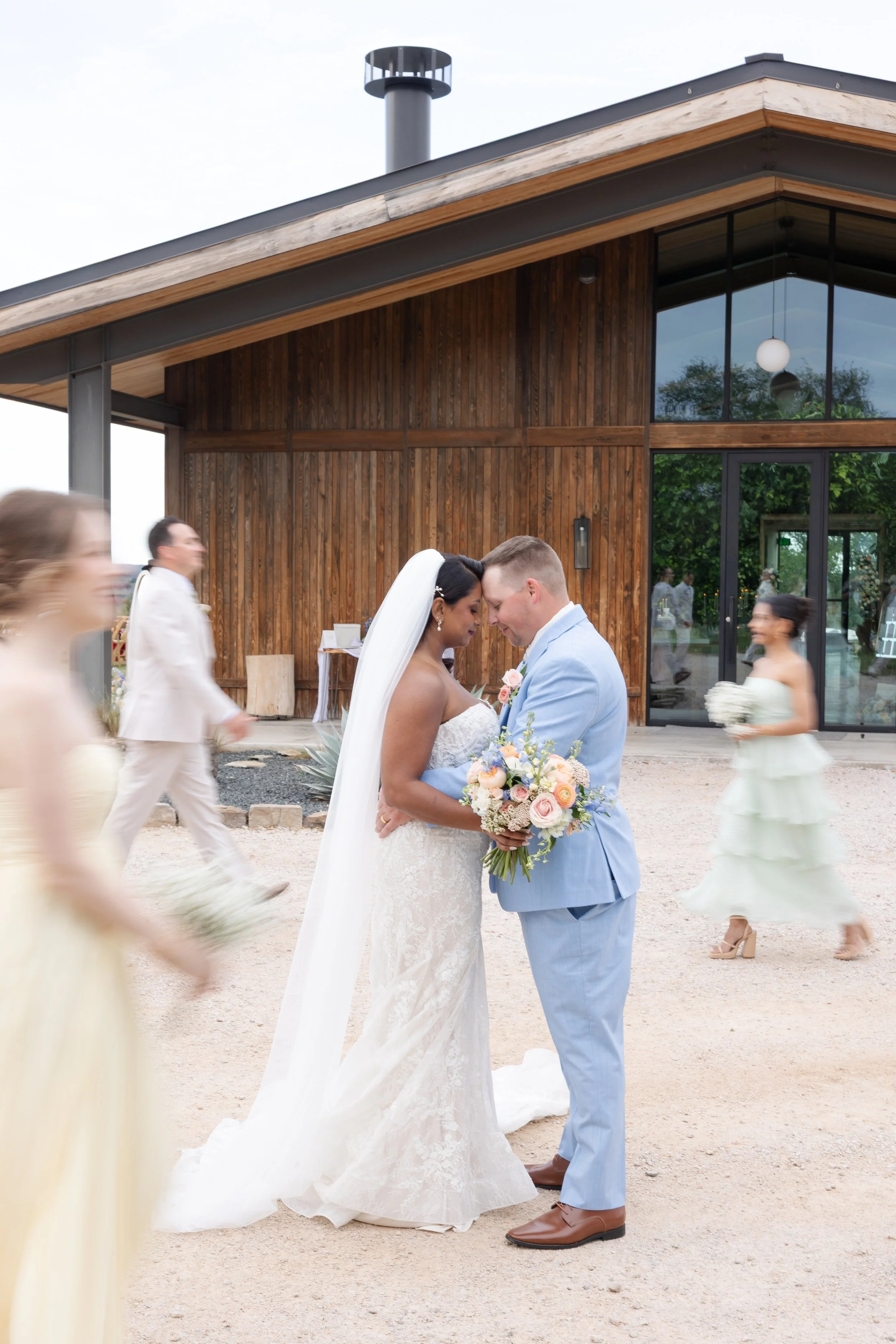 A bride and groom stand close together, touching foreheads, during their wedding ceremony outside. The bride wears a white lace wedding gown with a veil, and the groom wears a light blue suit with brown shoes. Bridesmaids and groomsmen, some blurred,