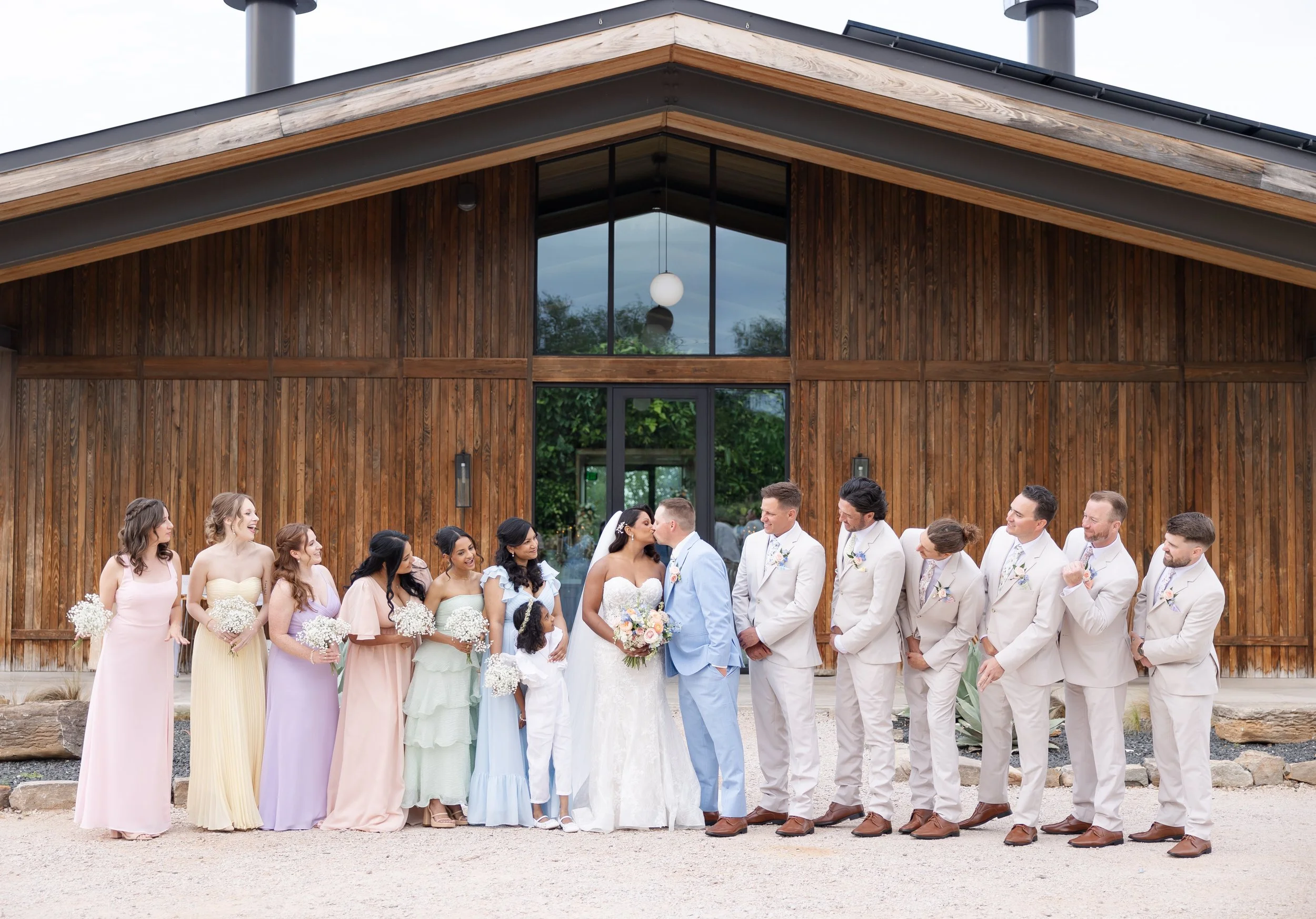 A group of people dressed in wedding attire standing in front of a modern wooden building with large glass doors, during  outdoor wedding party portraits at Walden Retreats.
