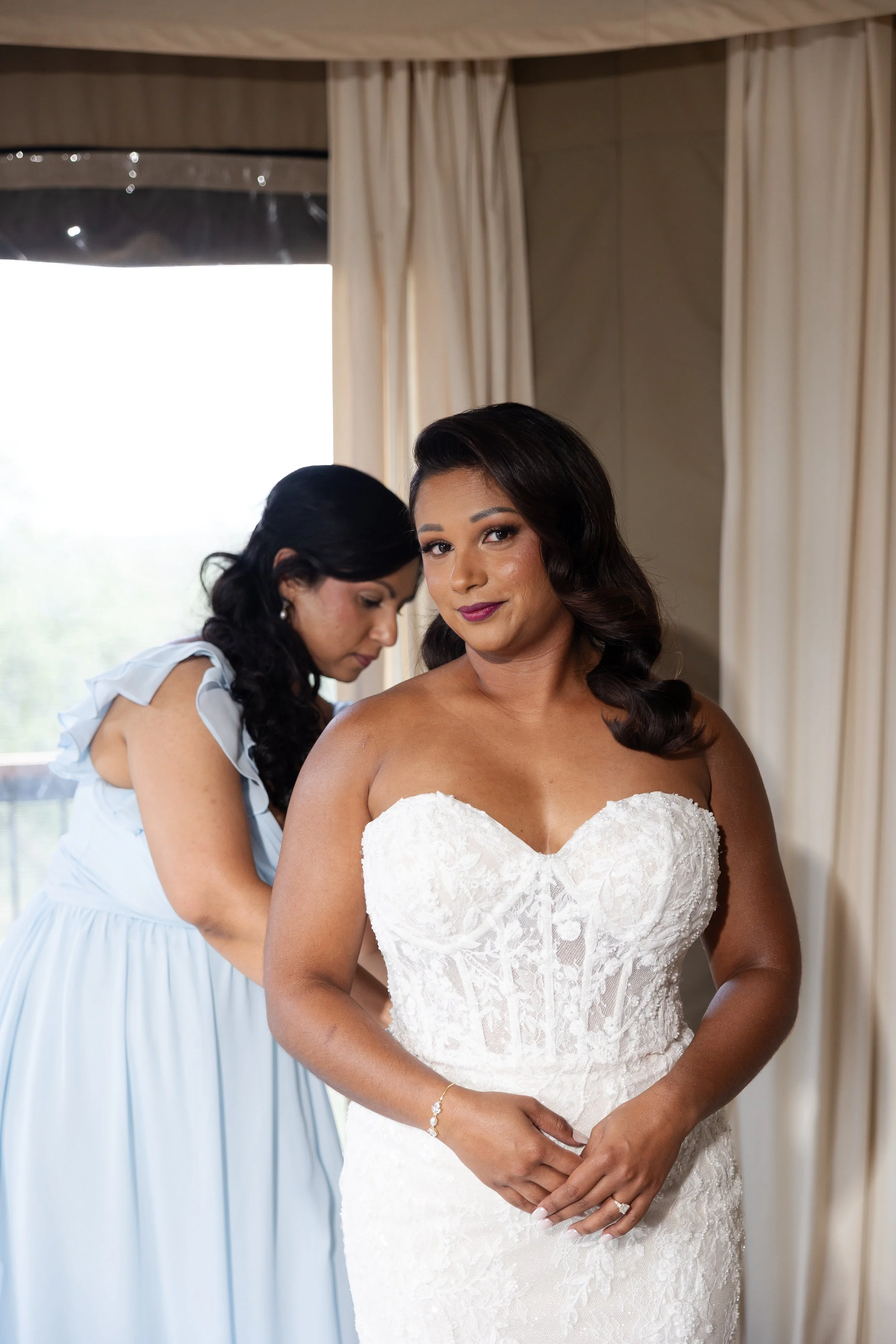 A bride in a strapless wedding gown stands with her hands clasped in front of her, while another woman helps her with her dress, indoors with beige curtains and a window in the background.