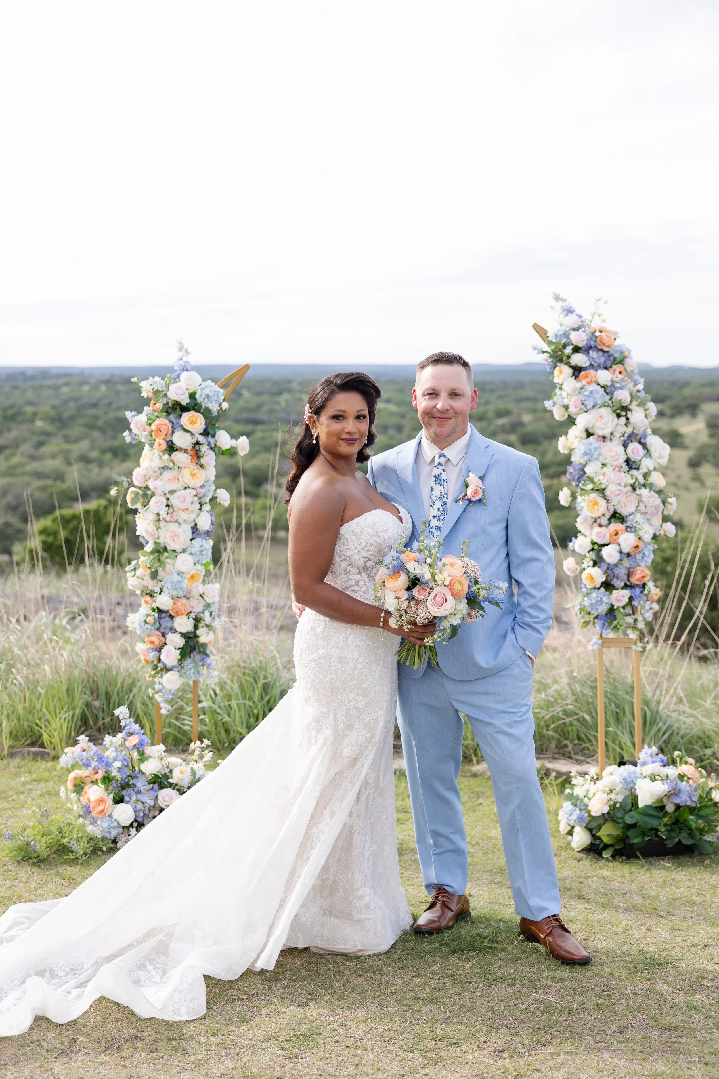 A newlywed couple standing outdoors on a cloudy day, with the bride wearing a white lace wedding dress and holding a bouquet of flowers, and the groom wearing a light blue suit and tie, with floral arrangements in the background at Walden Retreats.