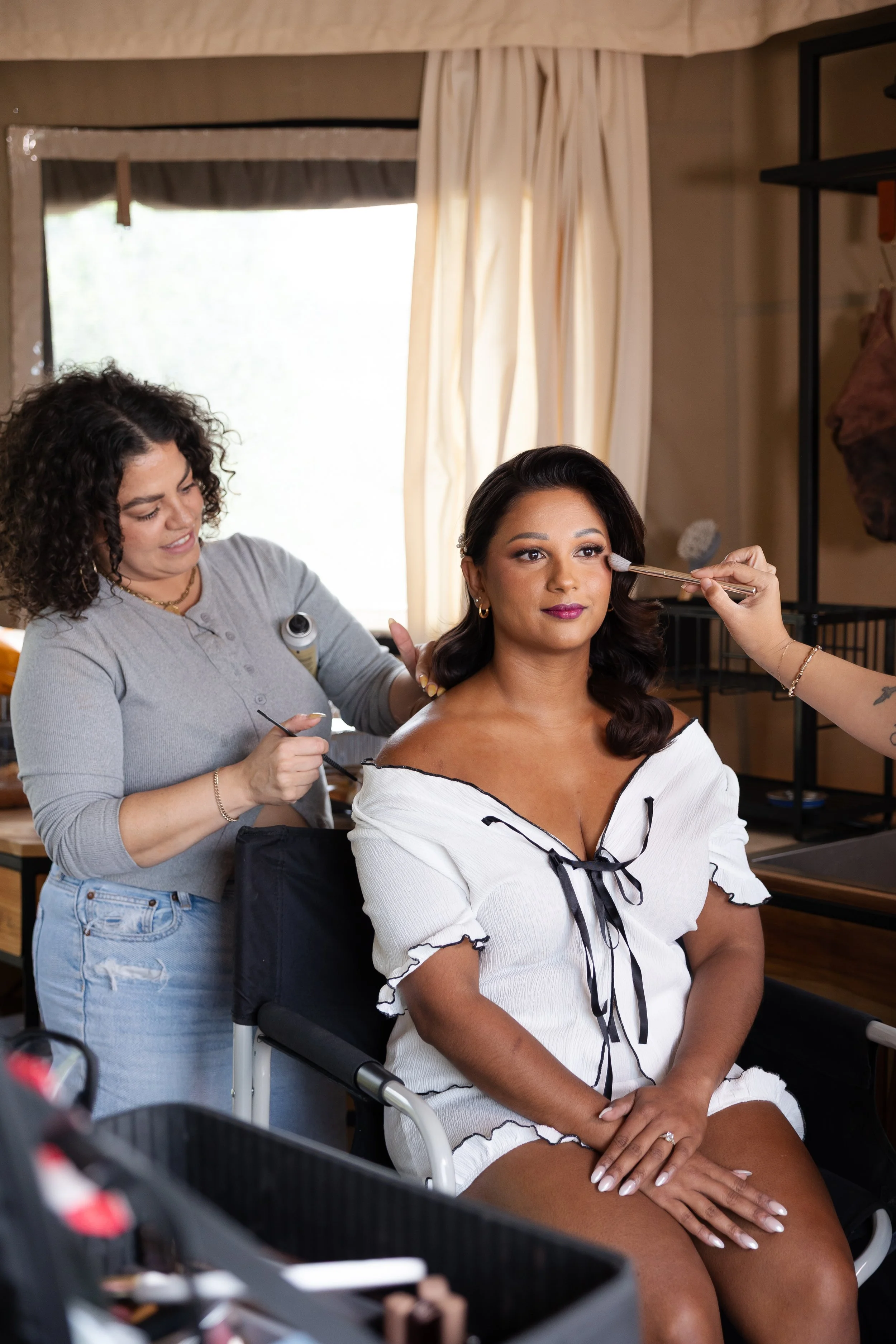 A woman with dark, curled hair sits in a makeup chair while a makeup artist applies eyeshadow. The woman is wearing a white robe with black accents and has her hands resting on her lap in the bridal suite at Walden Retreats.