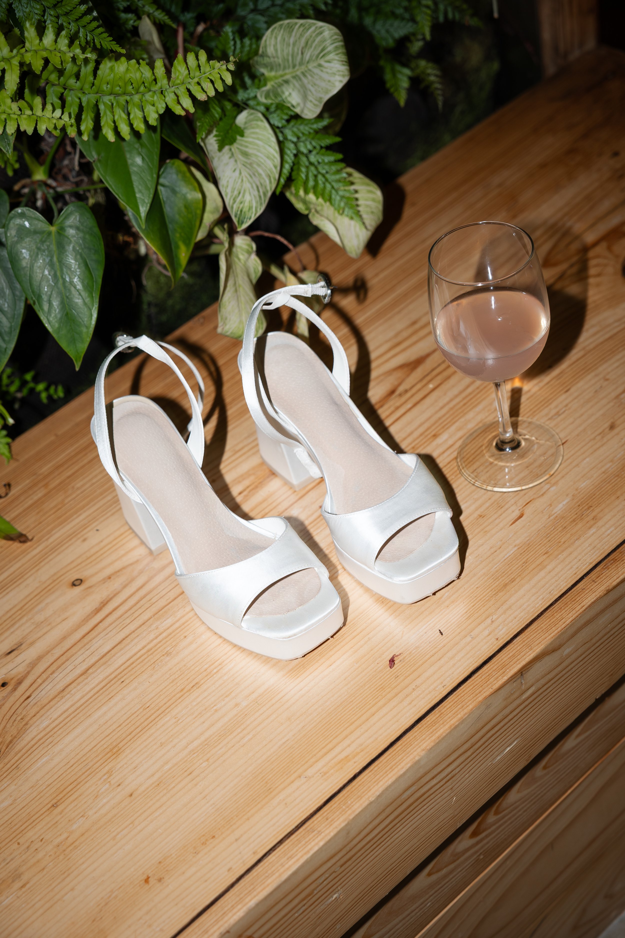 A pair of white high-heeled shoes and a glass of rosé wine on a wooden surface with green plants in the background.