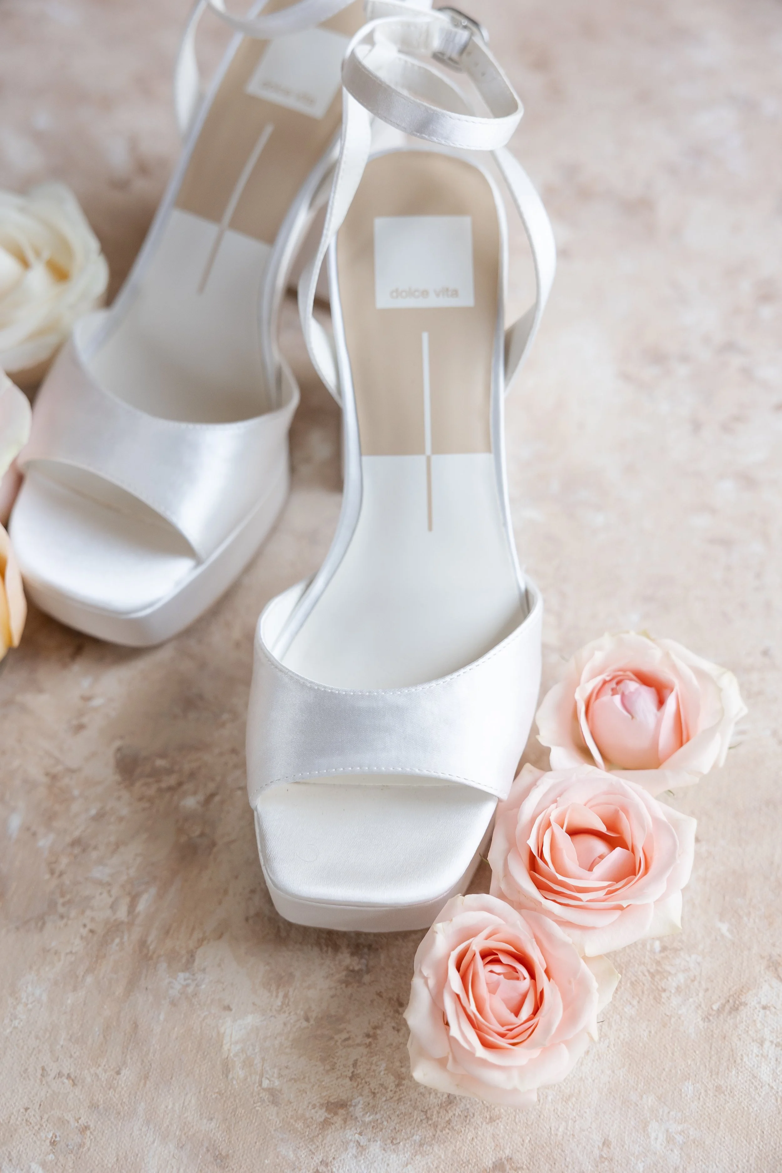 A pair of white high-heeled sandals with open toes and ankle straps, placed on a light-colored surface, surrounded by pale pink roses.