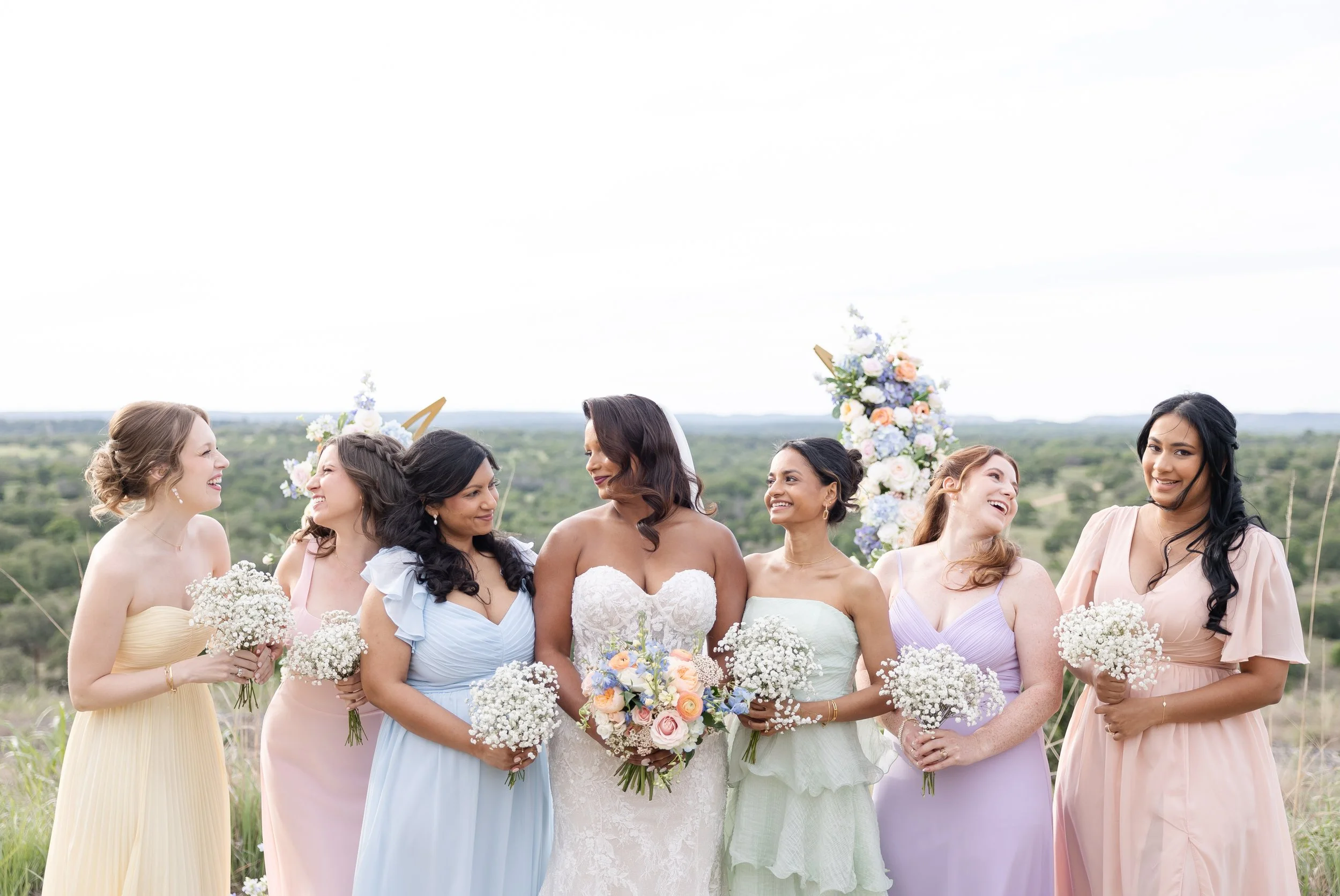 A group of bridesmaids and bride dressed in pastel-colored dresses, holding bouquets, standing outdoors with a floral arch and scenic landscape, at a wedding celebration at Walden Retreat.