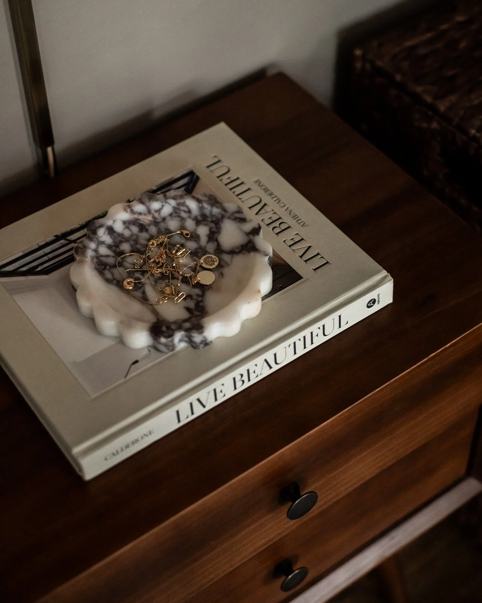 A marble-like jewelry dish with gold jewelry on it, placed on a large book titled 'Live Beautiful' on a wooden nightstand.