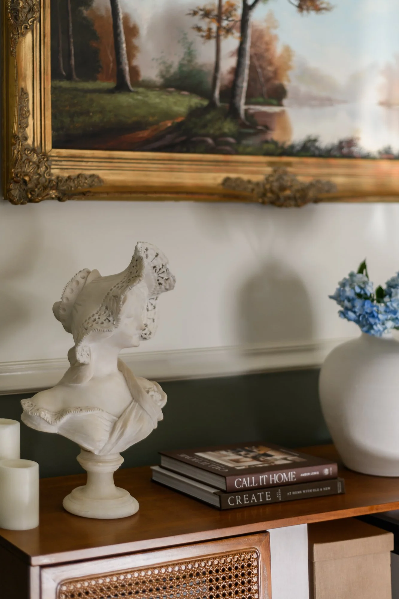 A corner of a wooden table holding a white bust sculpture, cylindrical candles, a stack of books titled 'CALL IT HOME' and 'CREATE', and a white vase with blue flowers. A landscape painting with trees and a river is hanging on the wall above the tabl