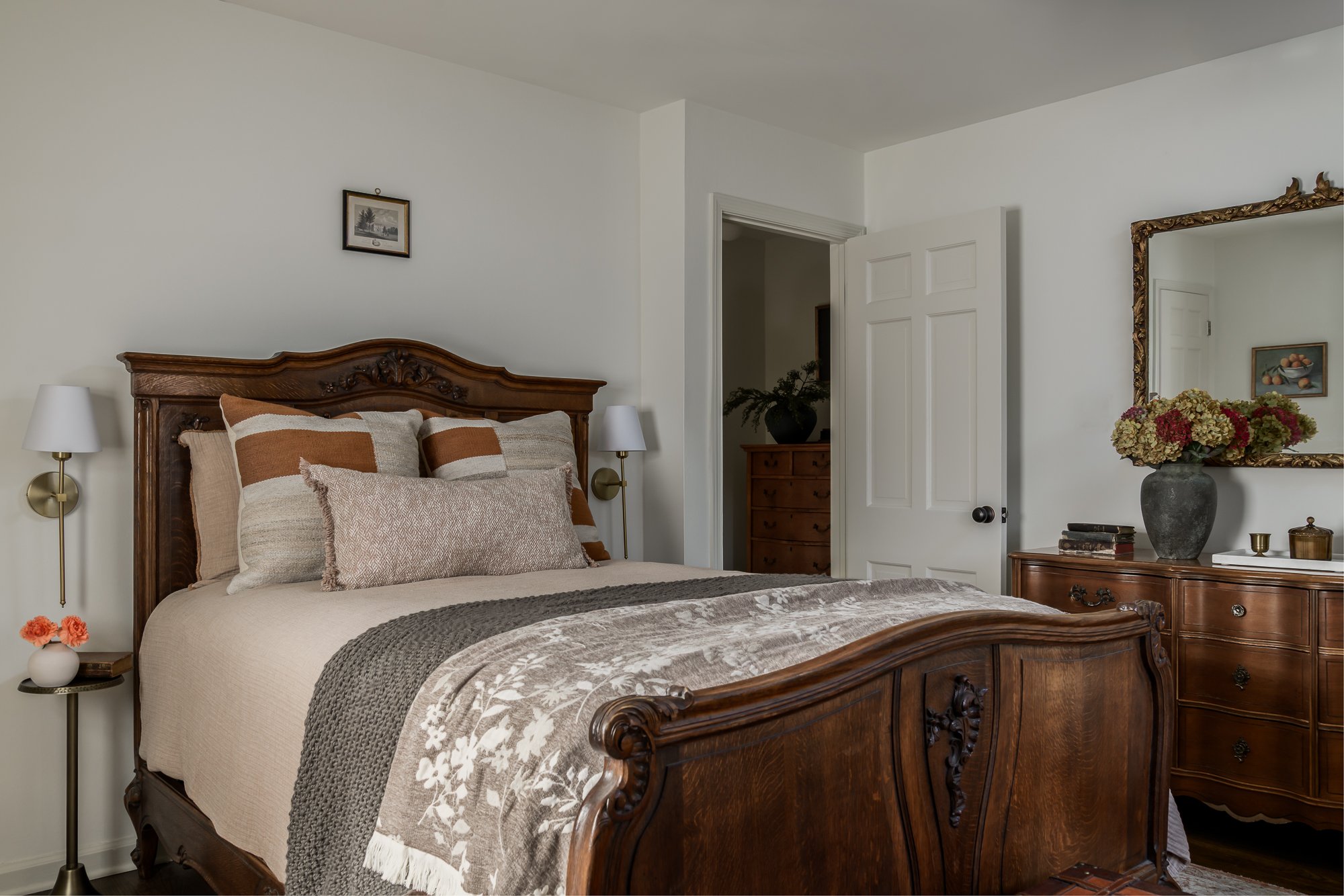 A bedroom with a wooden bed, beige bedding, decorative pillows, and matching bedside lamps. There is a dresser with a large mirror, a vase with flowers, and framed artwork on the walls.