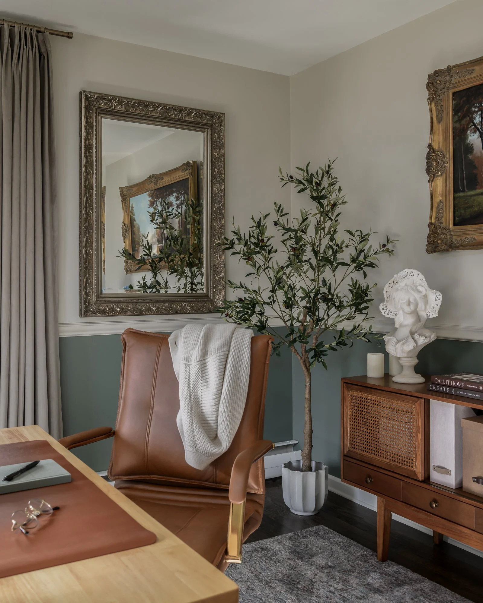 A home office with a large mirror, a potted tree, a wooden cabinet, a decorative bust, and a leather office chair with a white blanket draped over it.
