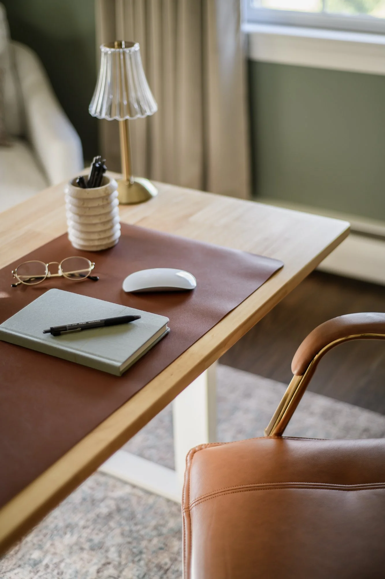 A wooden desk with a pink and beige desk mat, a closed notebook with a pen on top, glasses, a computer mouse, a ceramic holder with pens, a small lamp with a pleated lampshade, and a window with curtains.