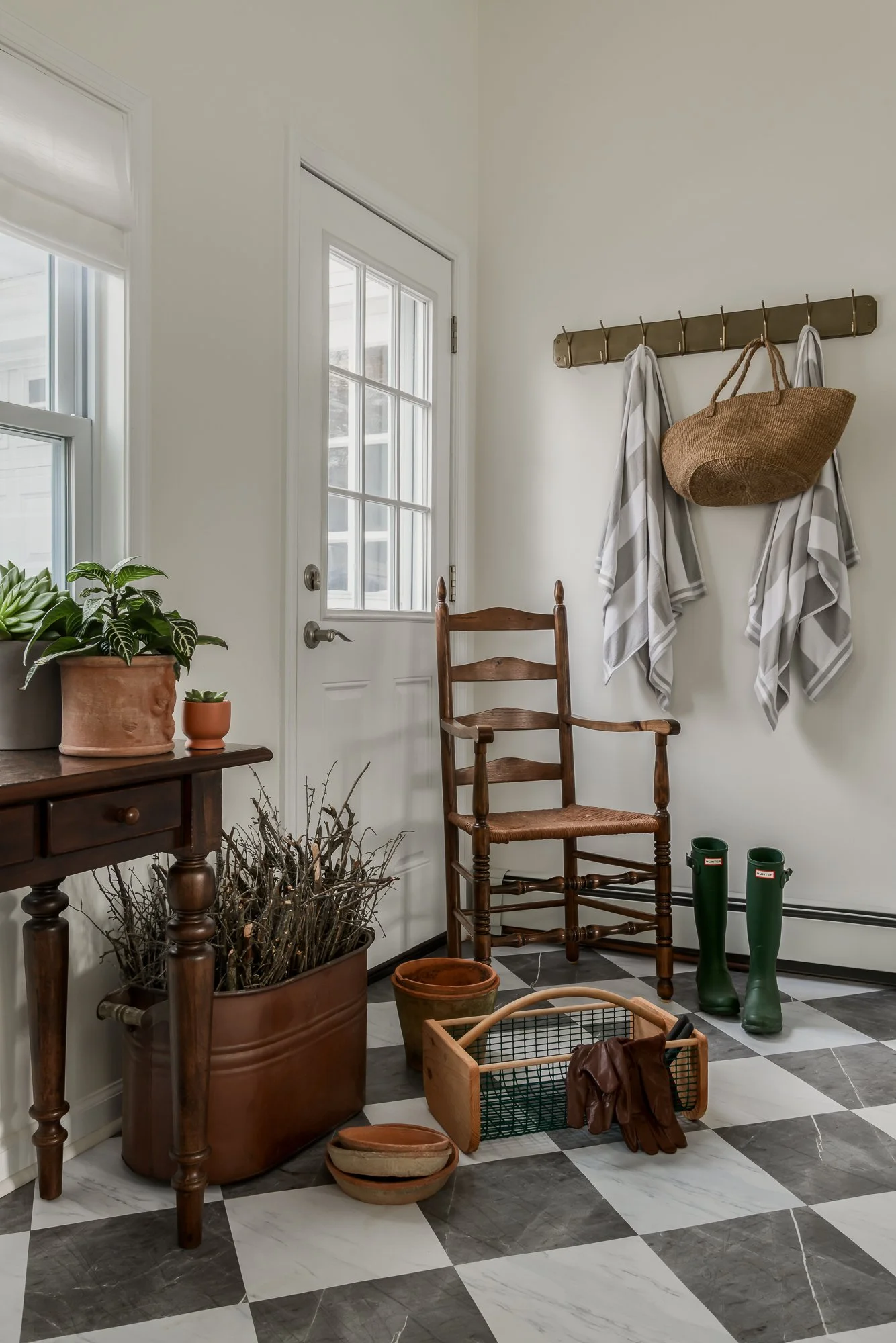 Entryway with checkered black and white tile floor, wooden chair, potted plants, coat rack with striped towels and a straw basket, green rain boots, and door with window.