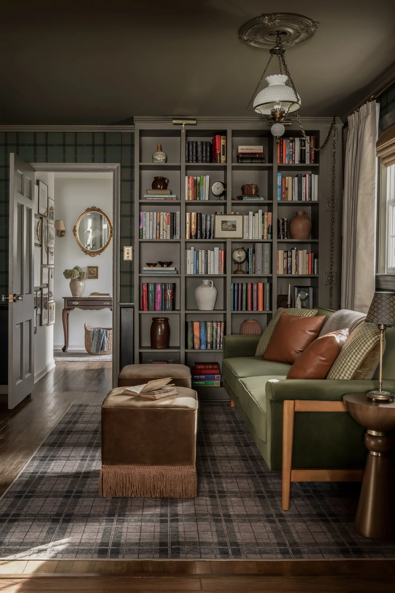 Living room with a green sofa, brown and plaid pillows, a wooden side table, a bookcase filled with books and decorative items, a window with curtains, and a ceiling light fixture.