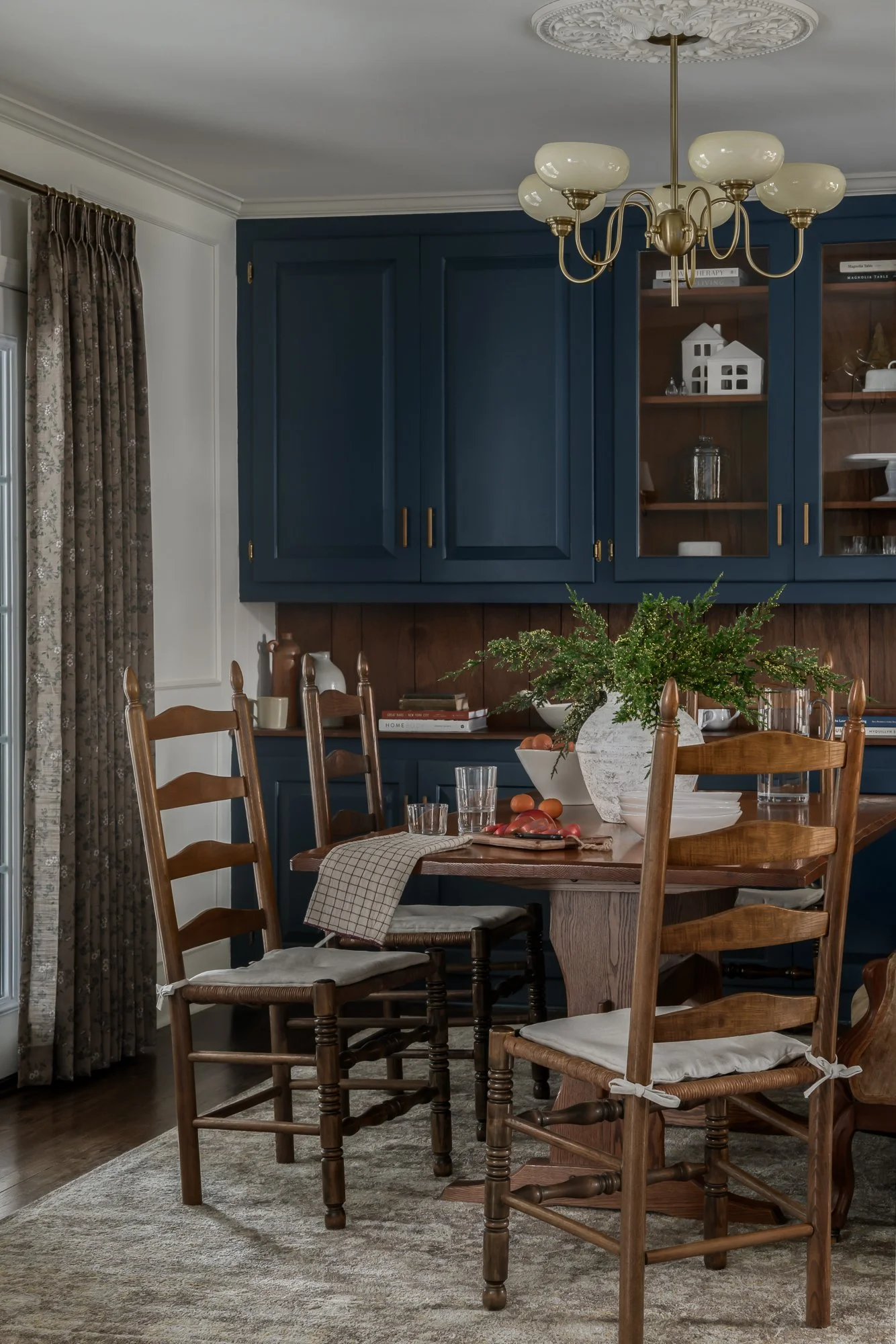 A dining room with a wooden table surrounded by six wooden chairs, a large ceramic vase with greenery, and a china cabinet painted in dark blue with open shelves and glass doors, decorated with books, glassware, and decorative items.
