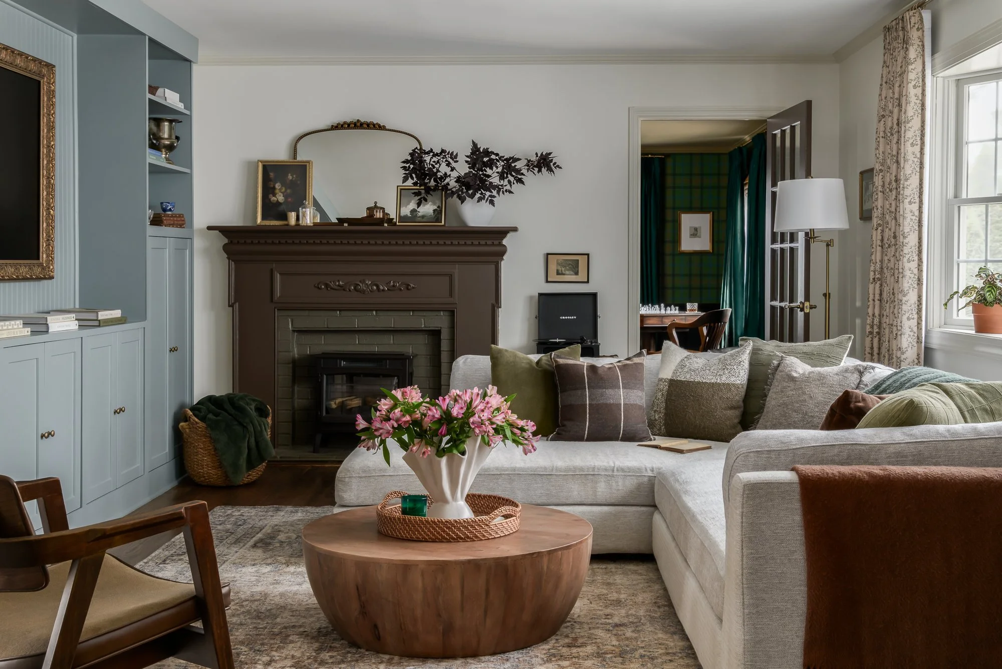 A cozy living room with a sectional sofa, a round wooden coffee table with a vase of pink flowers, a fireplace with a mantle, and a book and pillow on the sofa. The room has a large window with floral curtains, a blue built-in bookshelf, and a doorwa