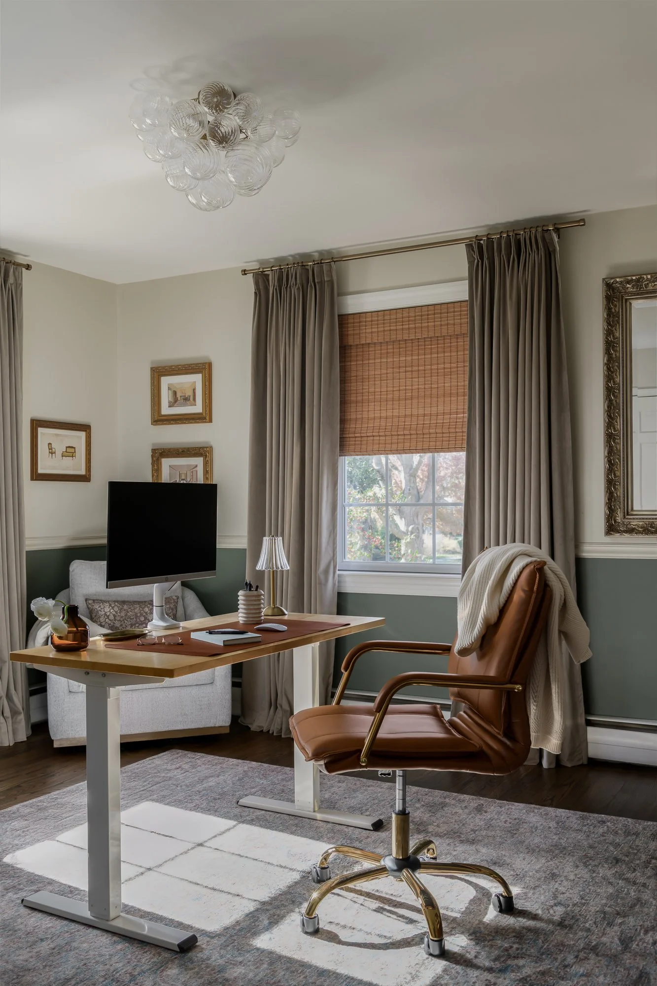 A home office with a wooden desk, brown swivel chair, computer monitor, small table lamp, and decorative objects, with beige curtains and framed artwork on the walls near a window with bamboo blinds.