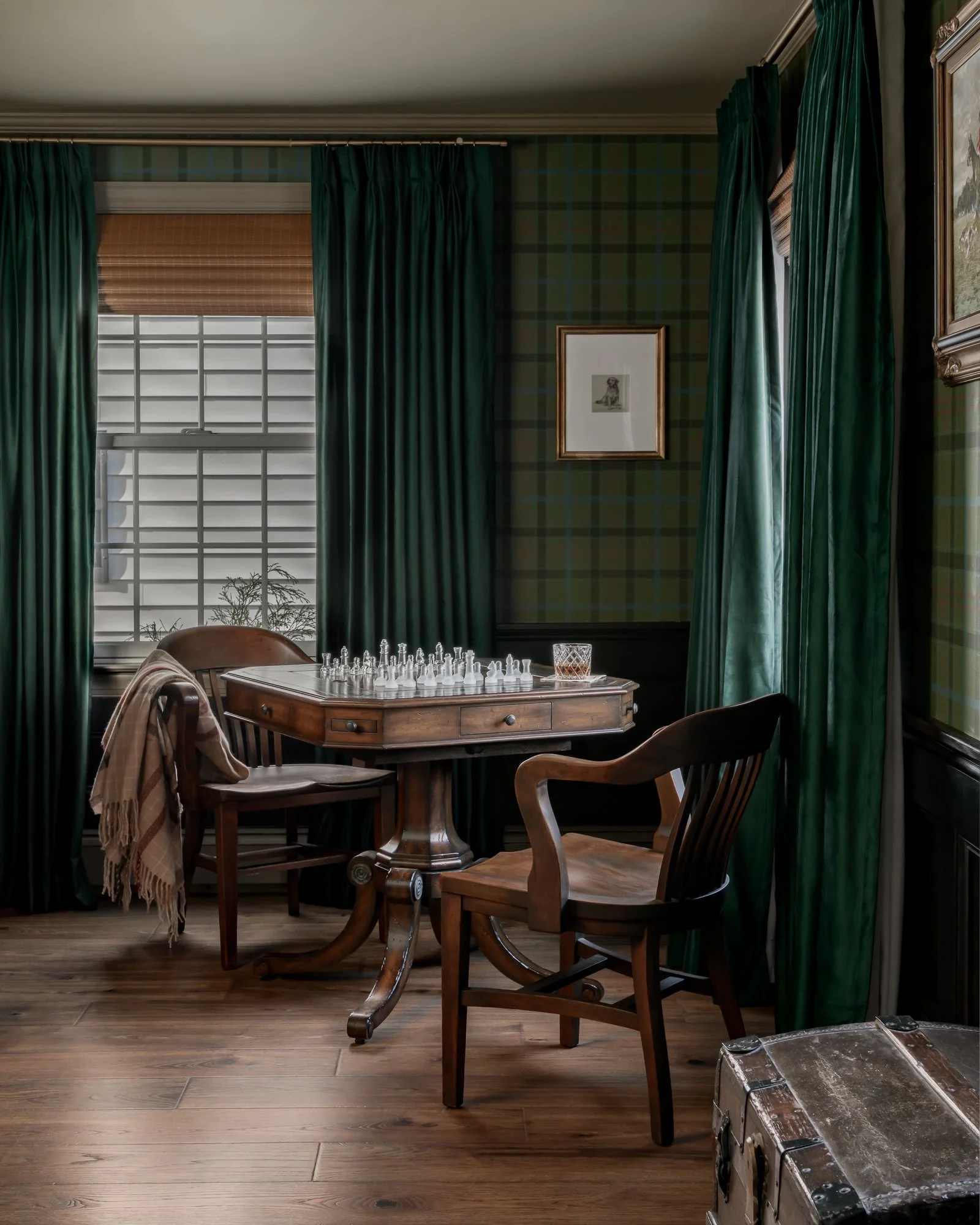 A cozy, vintage-style room with dark green curtains, plaid green wallpaper, a wooden table with a chess set, surrounded by wooden chairs, and framed artwork on the walls.
