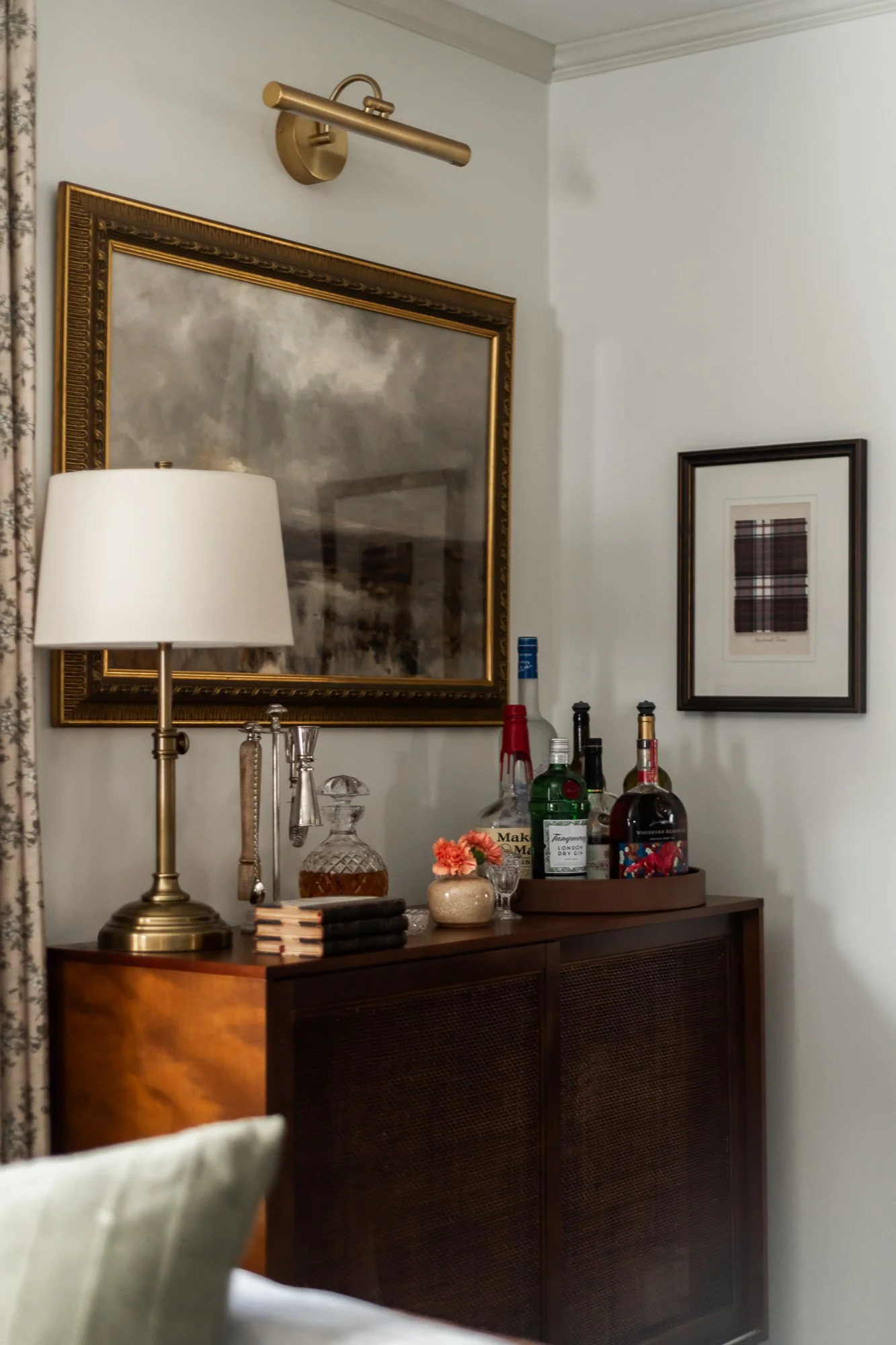 A wooden sideboard with a brass desk lamp, a decanter, small books, and a decorative vase with flowers, set against a wall with framed artwork and a large painting.