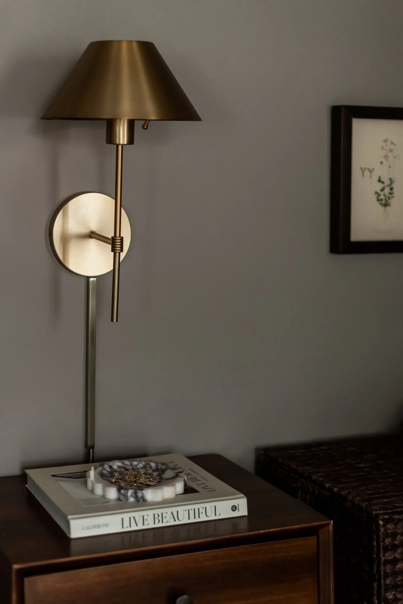 A wall-mounted gold and brass lamp next to a picture frame on a gray wall. Below, a wooden side table holds a stack of magazines, a white candle, and a decorative black and white marble tray with jewelry on top.