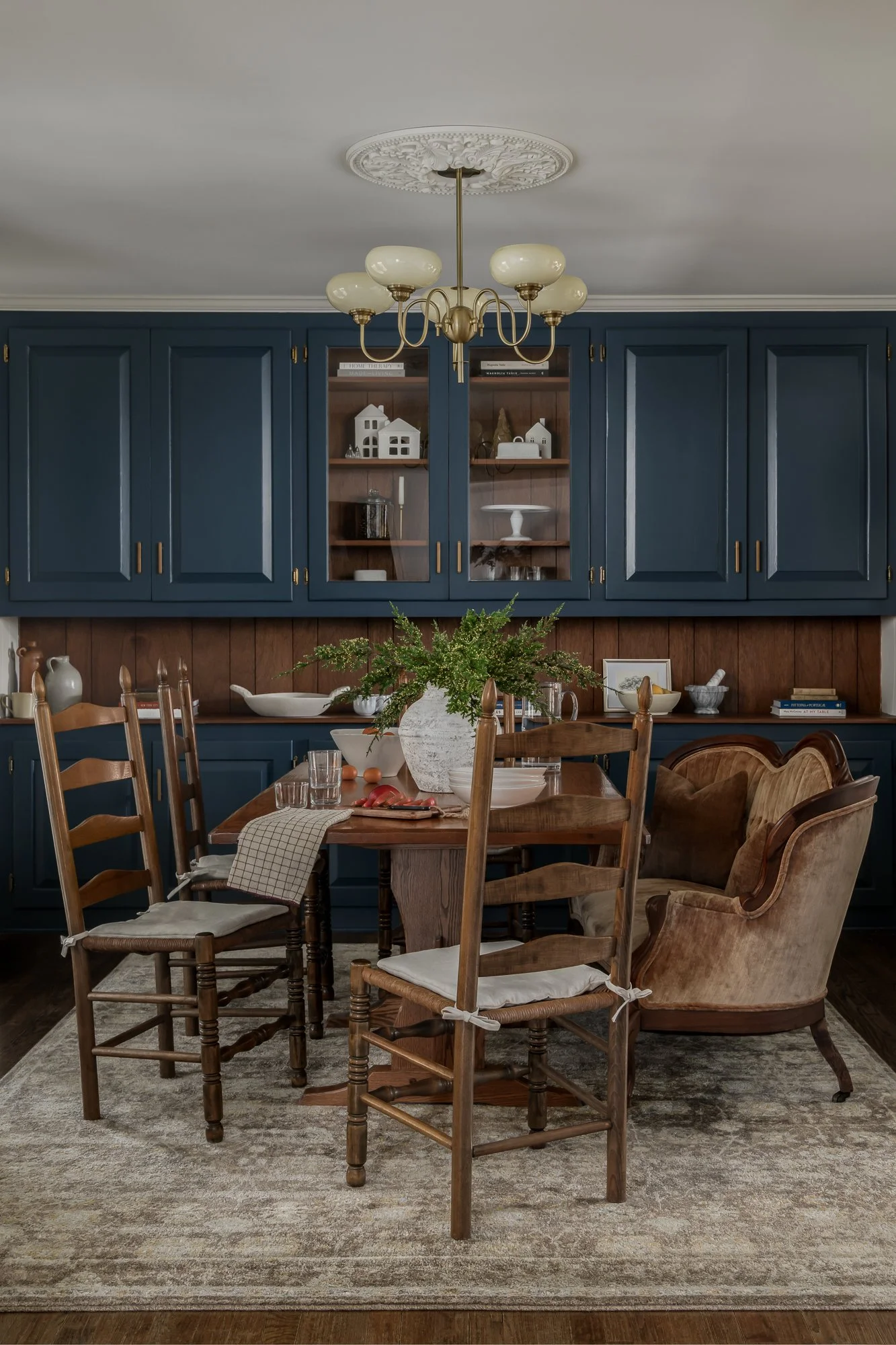 A dining room with a wooden table, six wooden chairs with padded seats, and a velvet armchair. A large green plant is in a white vase on the table. Blue cabinets with glass doors and wooden shelves are mounted on the wall behind the table. A chandeli