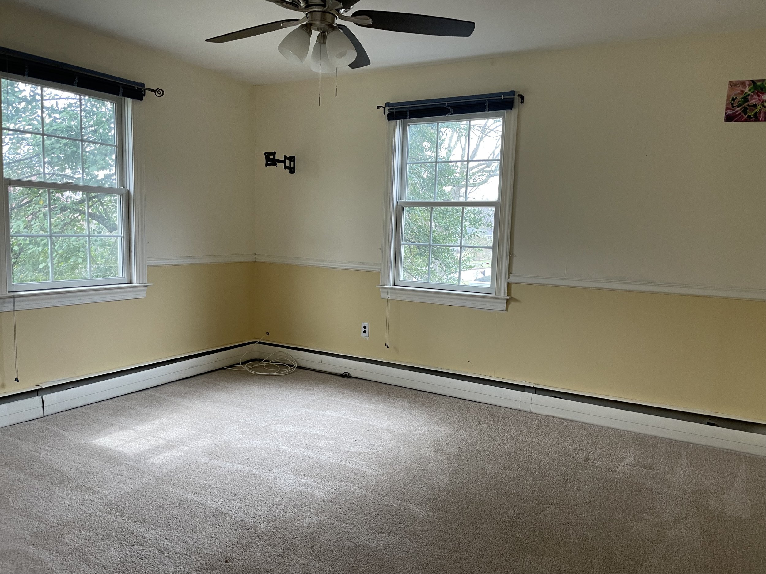 Empty room with beige carpet, yellow and white walls, two windows with blinds, a ceiling fan, and some cords on the floor.