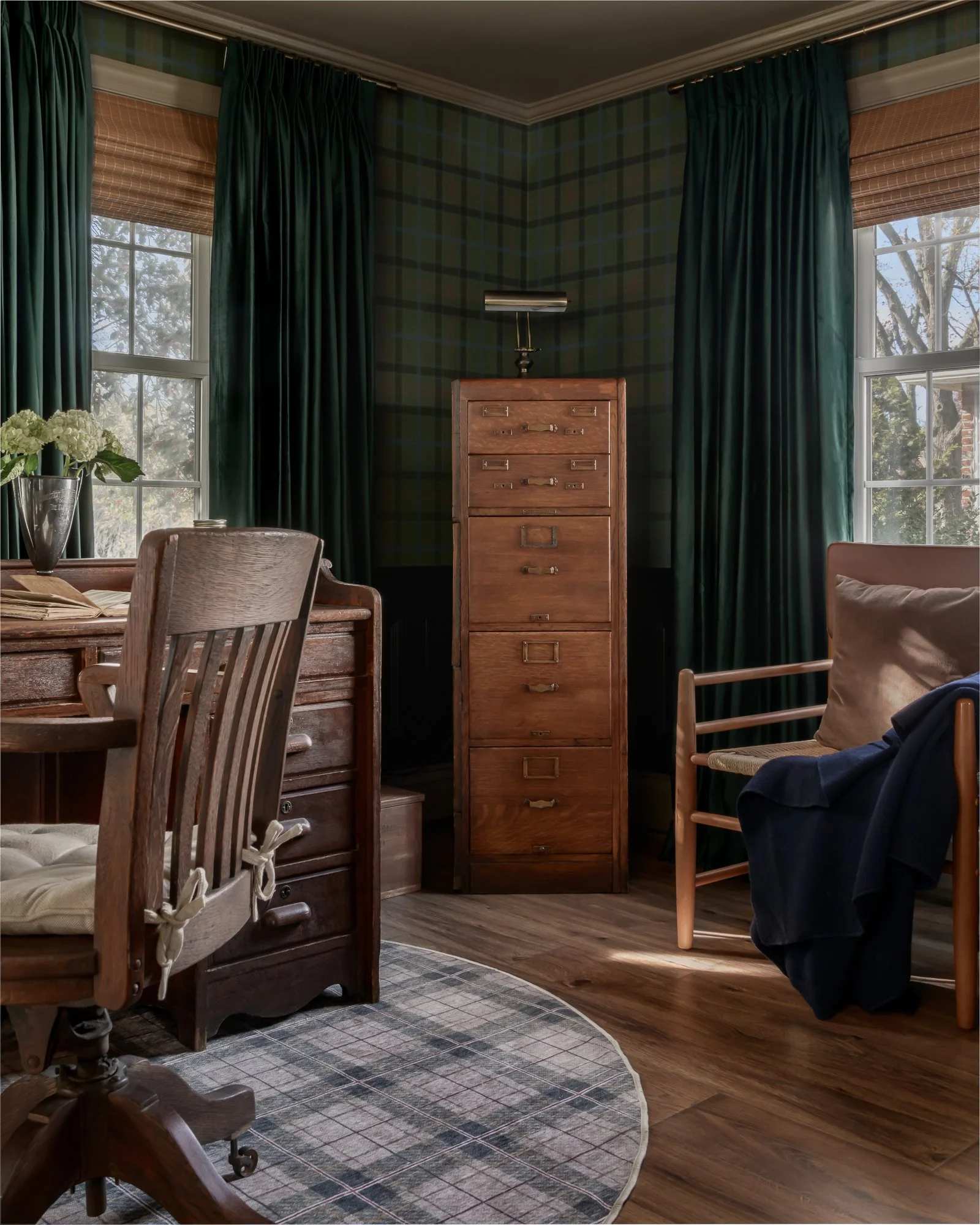 Interior of a room with green plaid wallpaper, dark green curtains, a wooden desk with a swivel chair with ribbon ties, a tall wooden filing cabinet, and two windows with views of trees outside.