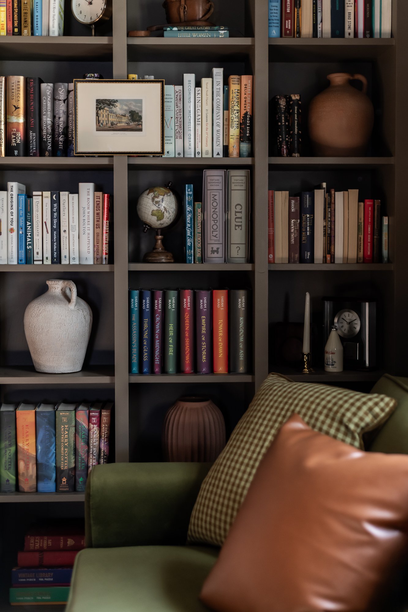 A bookshelf filled with books, decorative items, and a small globe. In front, part of a green armchair with two cushions, one checkered and one leather.