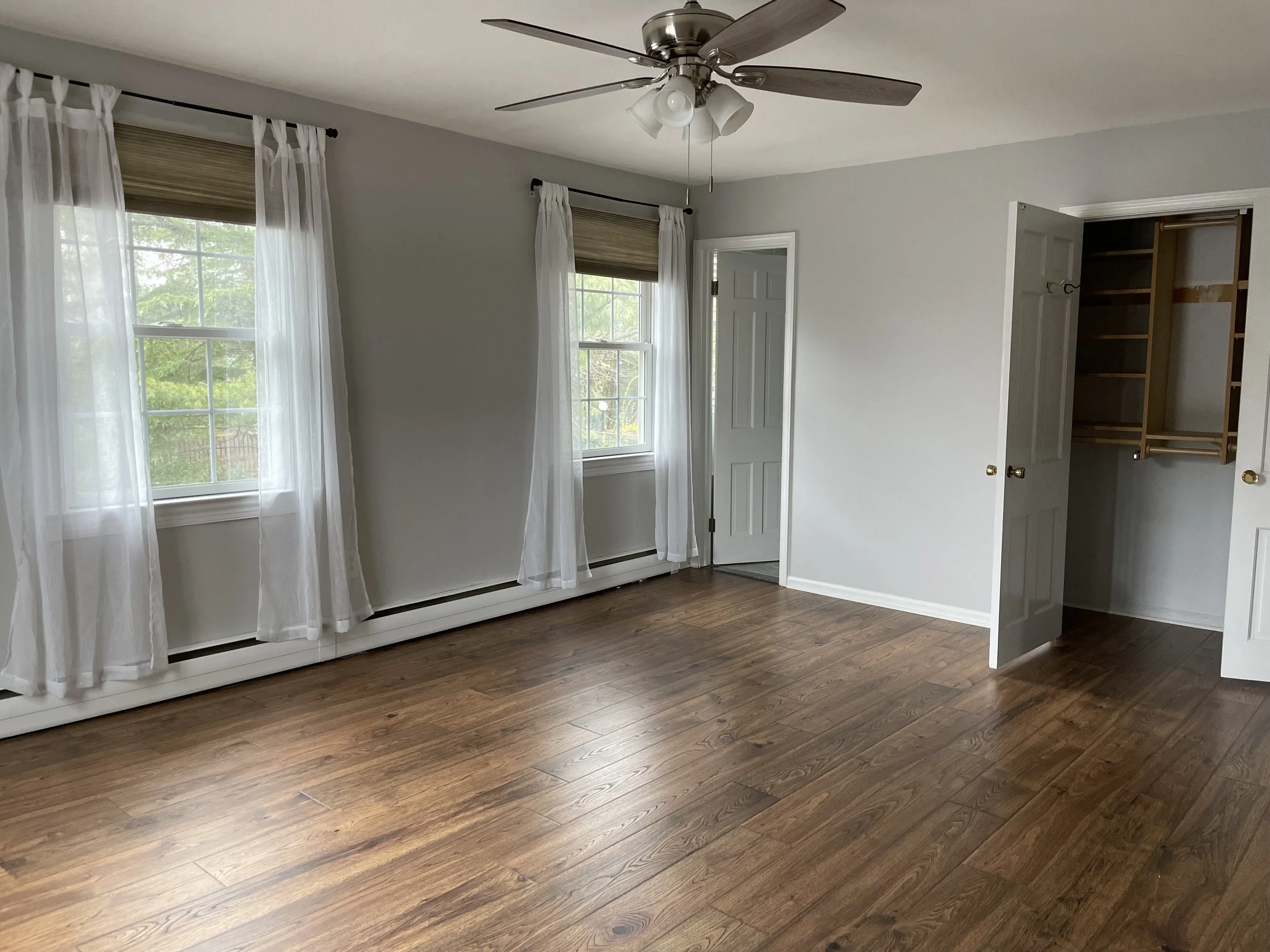 Empty room with hardwood floors, white walls, three windows with sheer white curtains, a ceiling fan, an open closet with wooden shelves and hanging space, and a door.