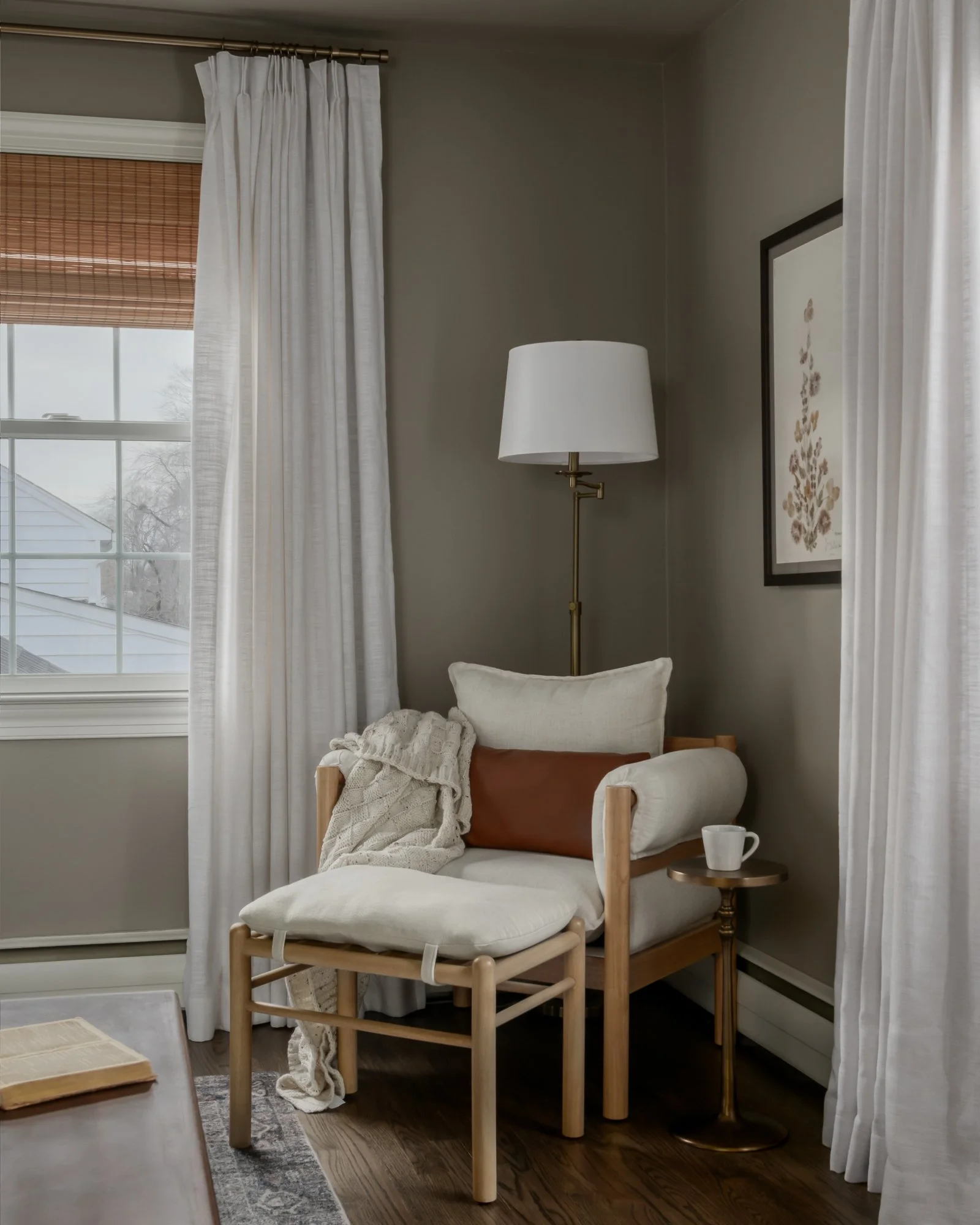 Cozy corner with a wooden armchair draped with a cream cushion, a brown bolster pillow, a Bench with a cushion, a side table with a mug, a floor lamp, white curtains, and a framed botanical print on a gray wall, near a window showing a snowy outdoors