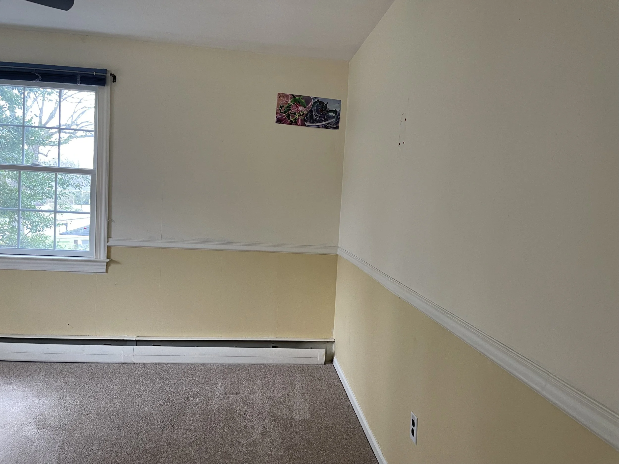 Empty room with beige carpeted floor, yellow half-wall and white wall with a window showing trees outside, a small colorful picture on the wall, and an electrical outlet.