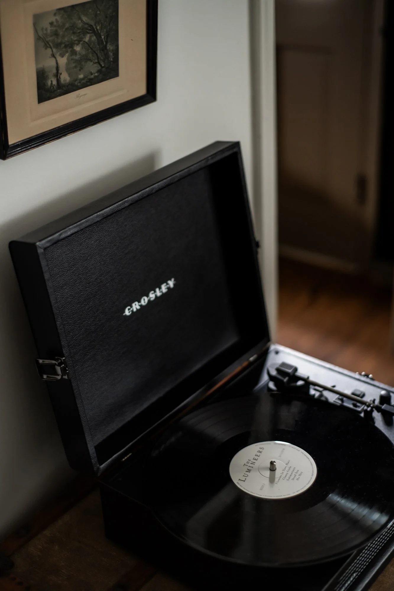 A vintage Crosley record player playing a vinyl record, situated on a wooden surface near a wall with a framed black-and-white landscape picture hanging above.